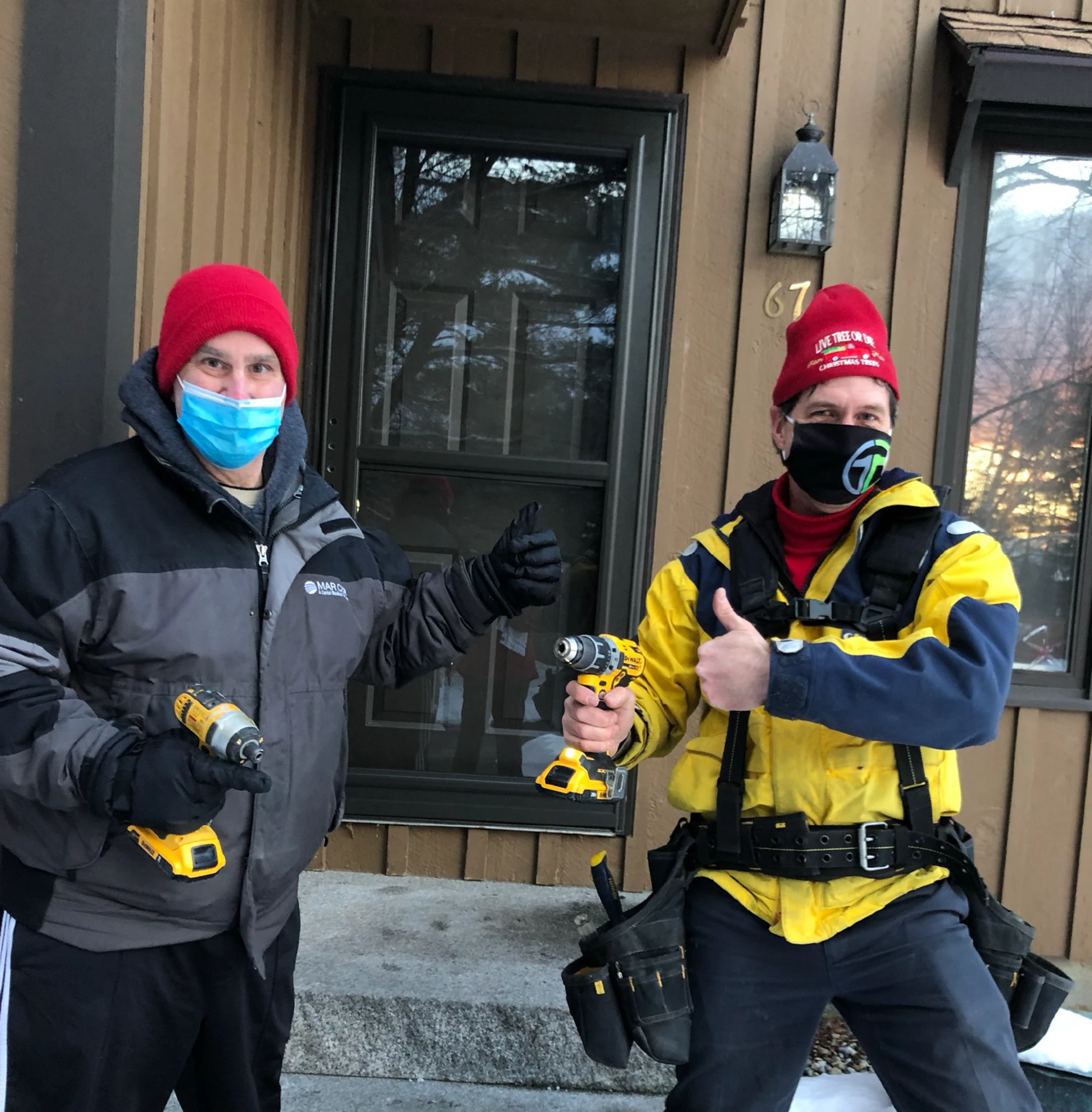 Two men in winter jackets and face masks stand in front of a house. They are both holding power drills and giving a thumbs-up gesture to the camera.