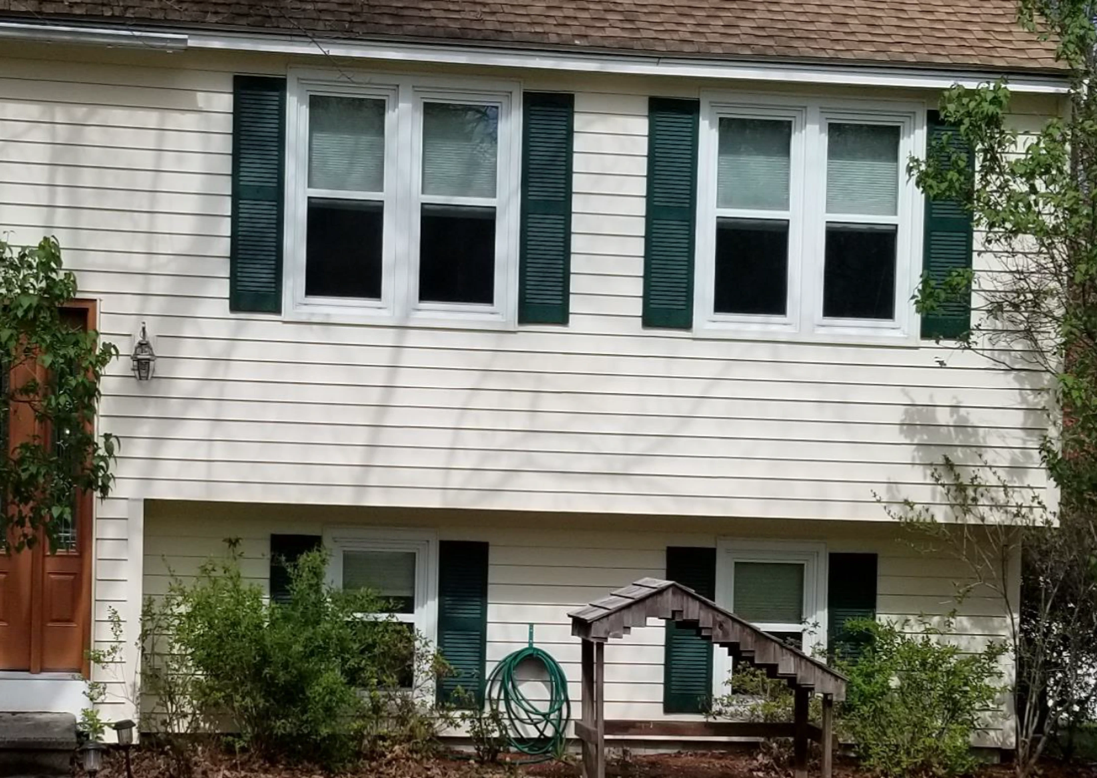 The front of a two-story home with light-colored siding, white-framed windows, and dark green shutters. Bushes and trees grow around the front of the house.