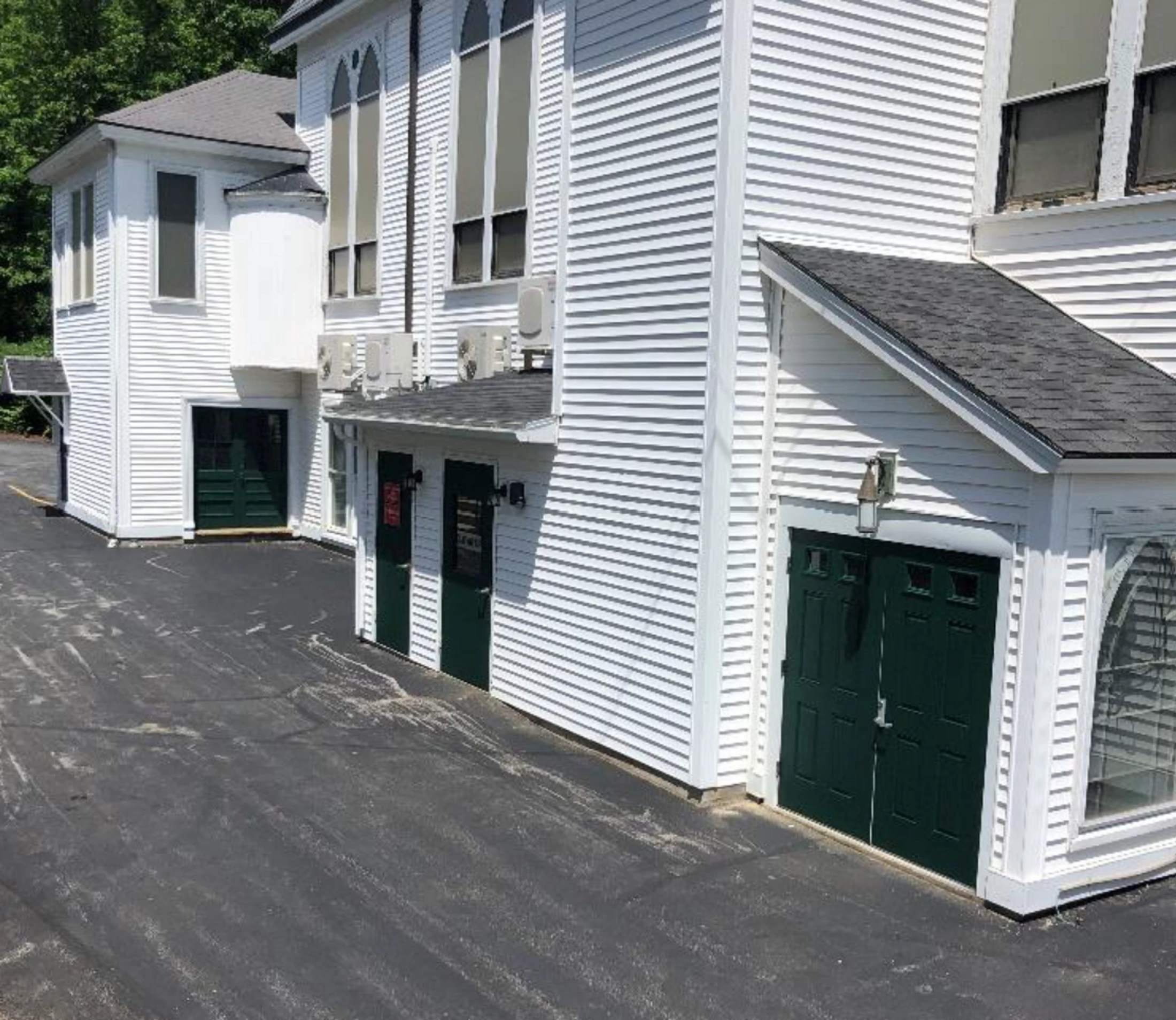 The side of a large building with white siding and multiple dark green doors, next to a black asphalt driveway on a sunny day.