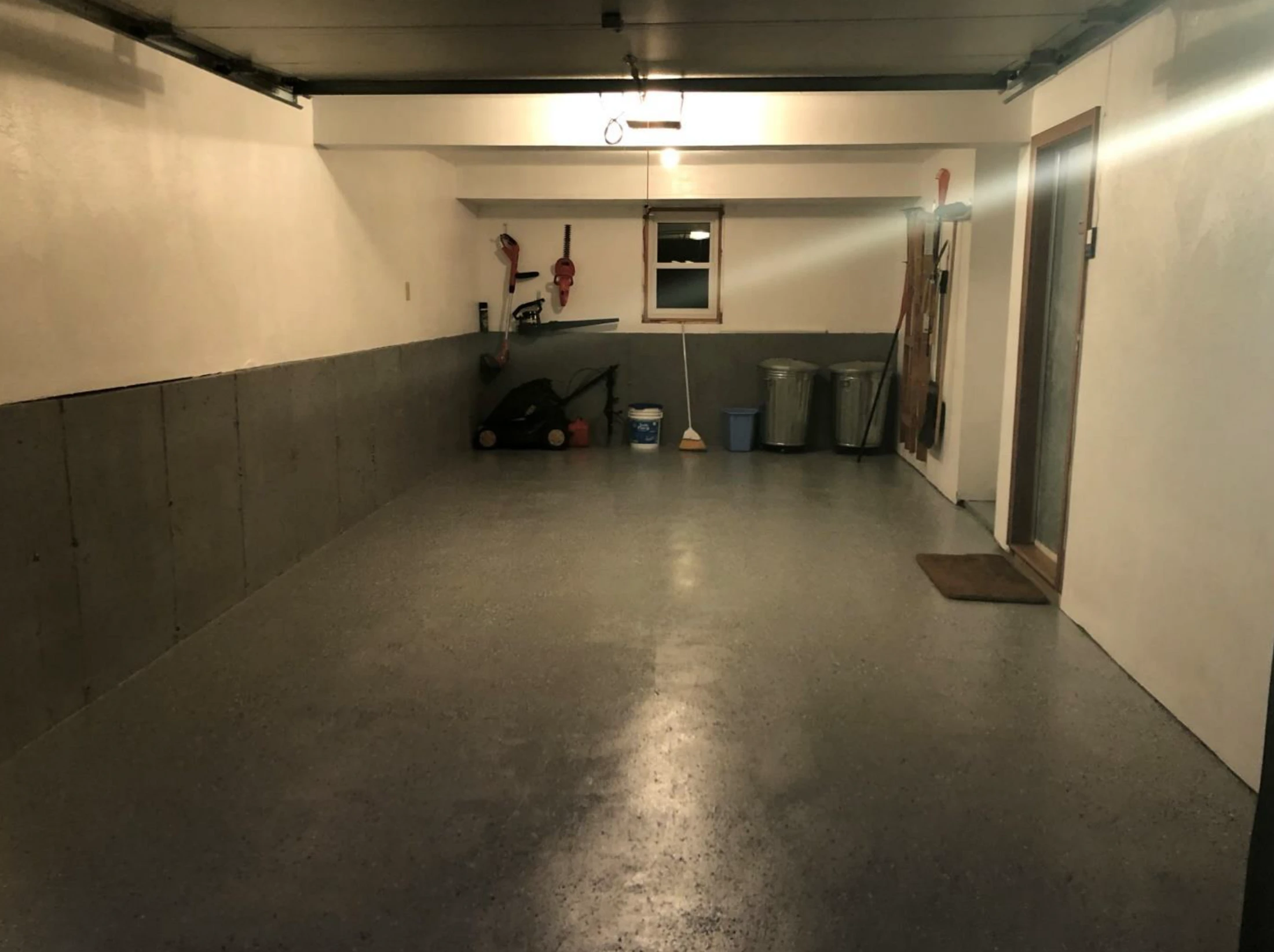A wide view of a clean, well-lit garage with a glossy gray speckled floor, white walls, and neatly stored lawn equipment along the back wall.