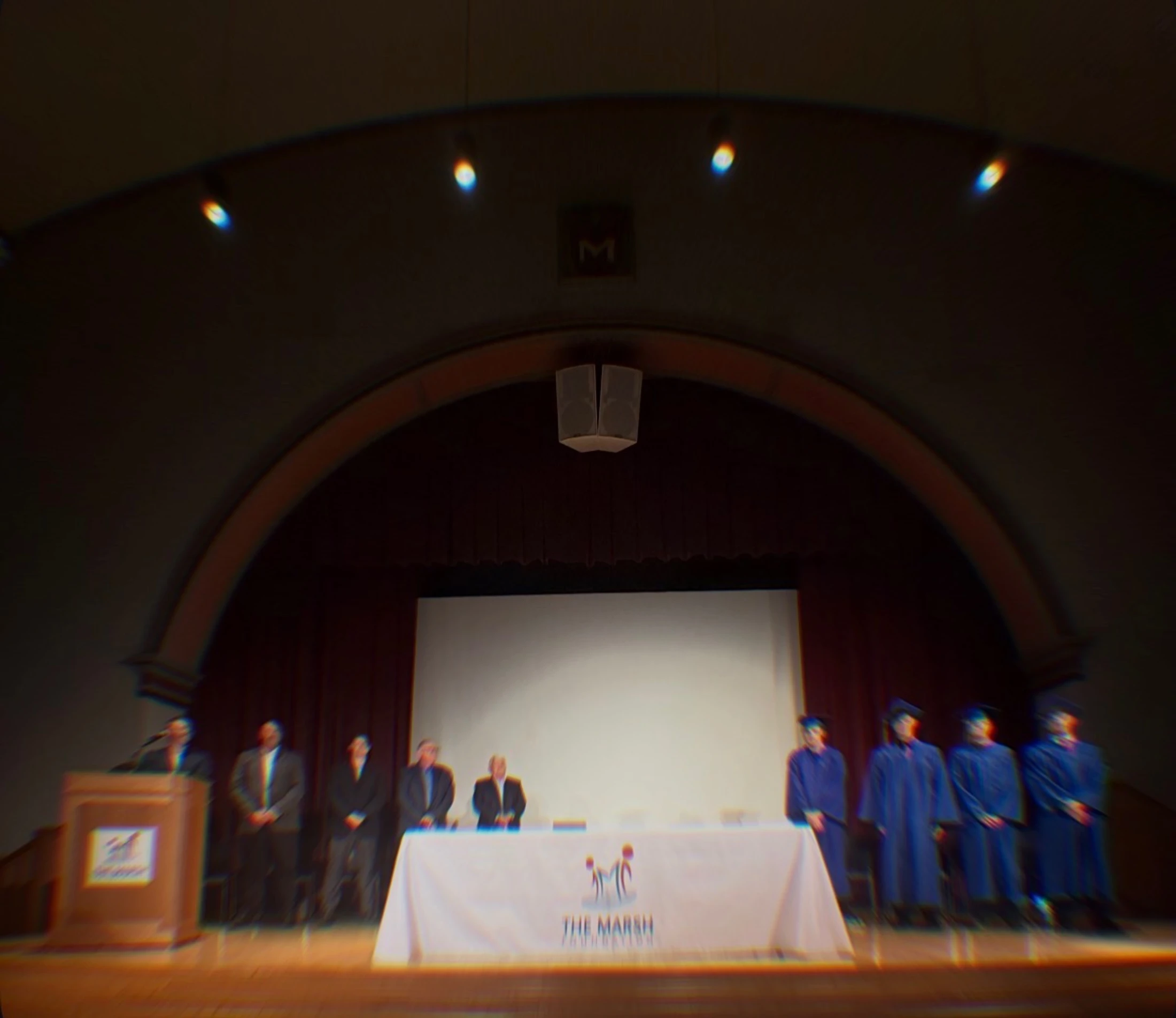 A blurry, dreamlike photo of a graduation-style ceremony on an auditorium stage with people in caps and gowns and others in suits.