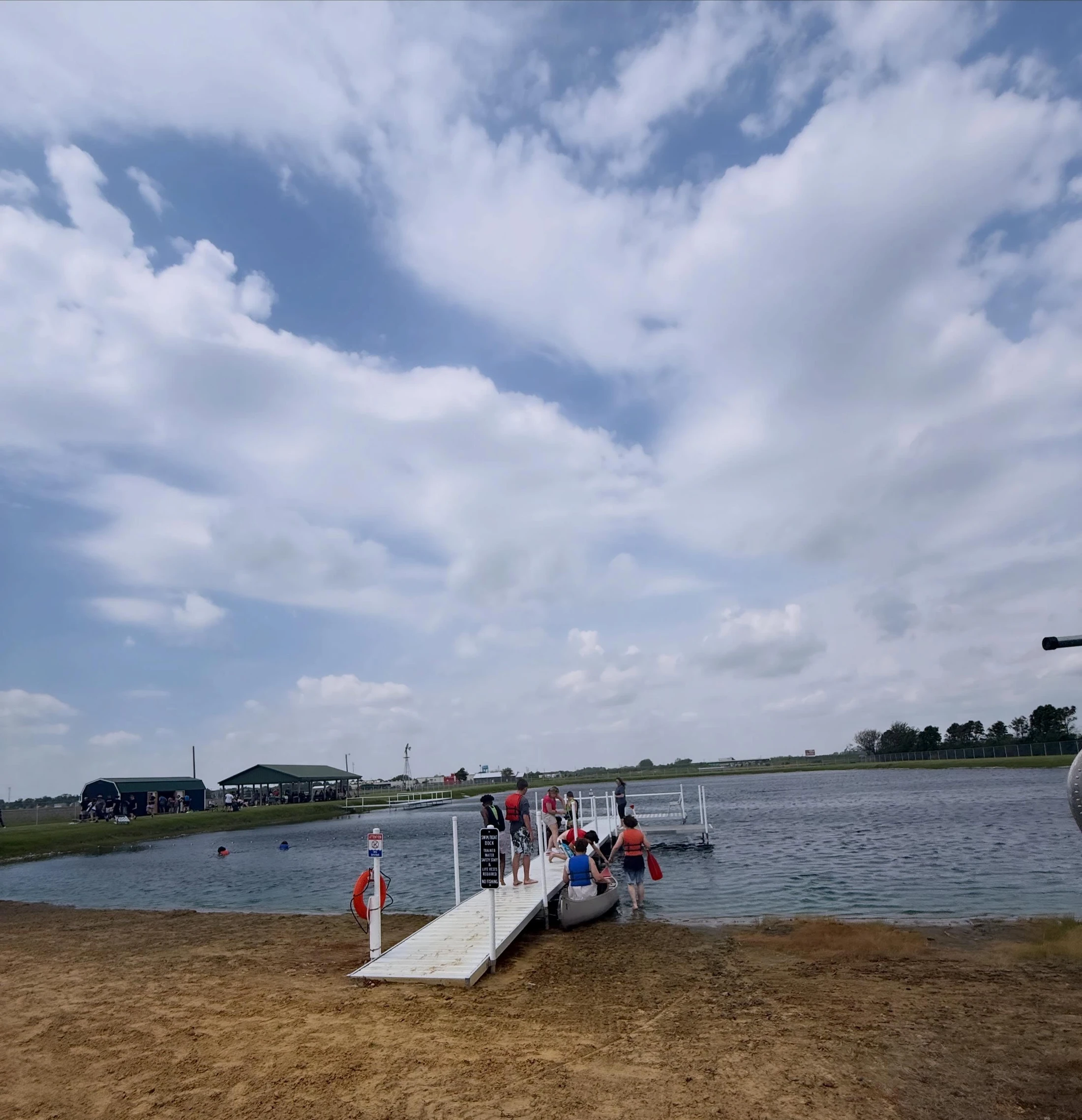 A group of people in life jackets gather on a white dock at a lake's sandy shore, preparing to get into a canoe under a partly cloudy sky.