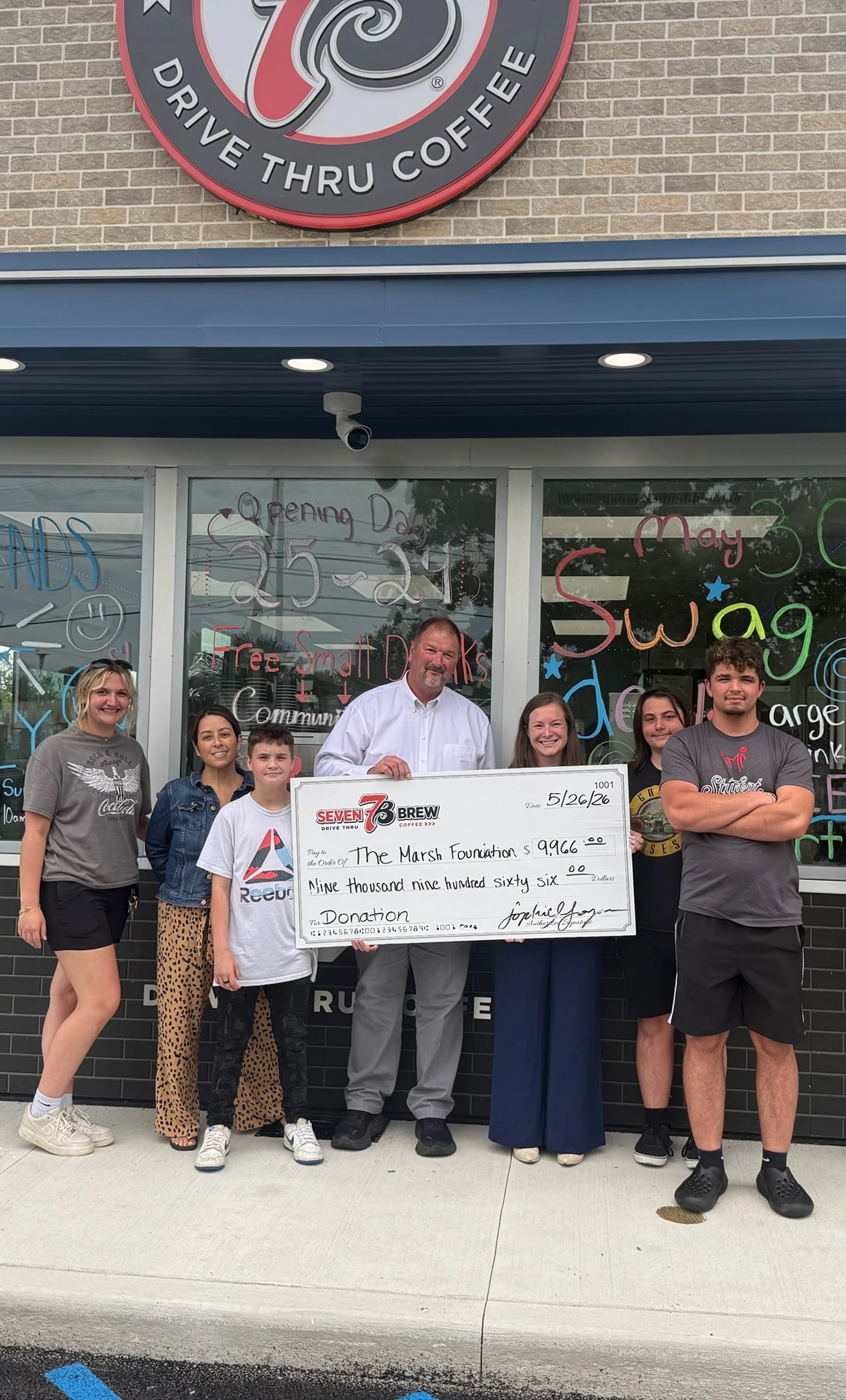 A group of people stand outside a 7 Brew coffee shop holding a large donation check for $9,966 payable to The Marsh Foundation.
