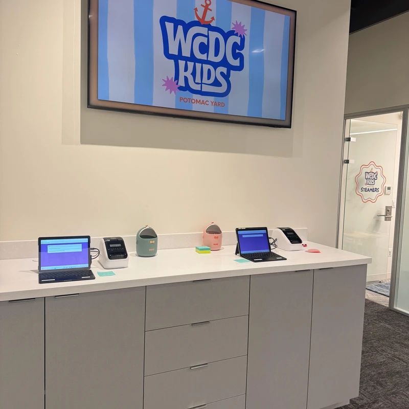 A modern check-in area with laptops and label printers on a counter. A large screen on the wall displays the 'WCDC KIDS Potomac Yard' logo.