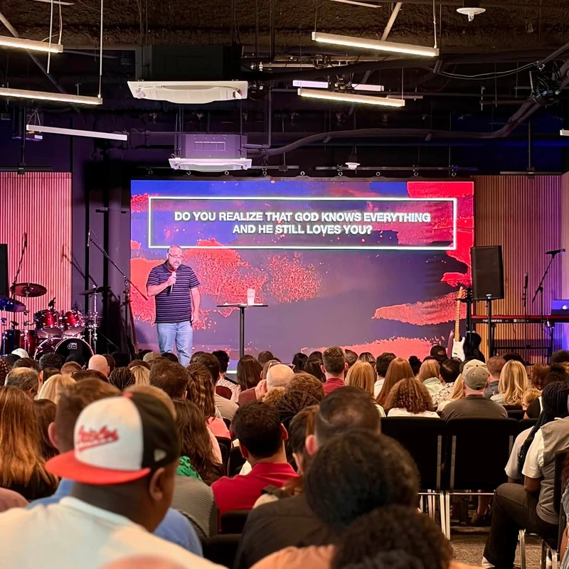 A pastor speaks on stage to a seated audience. Behind him, a large screen displays the words "DO YOU REALIZE THAT GOD KNOWS EVERYTHING AND HE STILL LOVES YOU?".