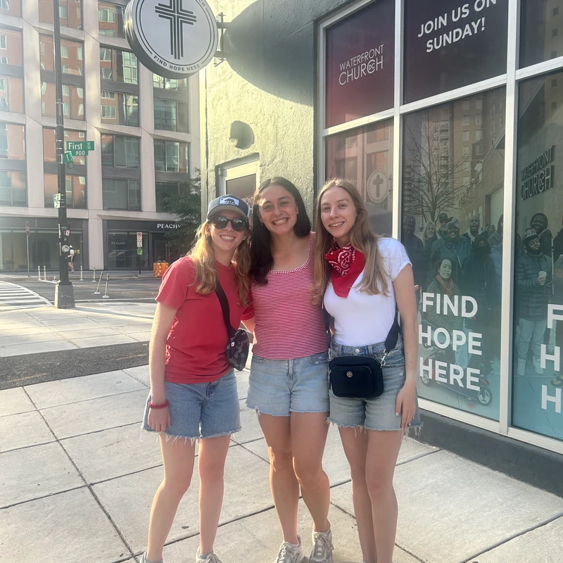 Three young women smile while standing on a city sidewalk in front of the Waterfront Church on a sunny day.