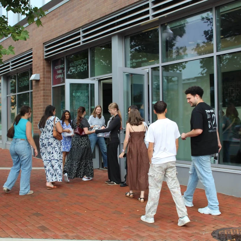 A group of young adults gather on a brick sidewalk outside the glass entrance to the Waterfront Church, a modern brick building.