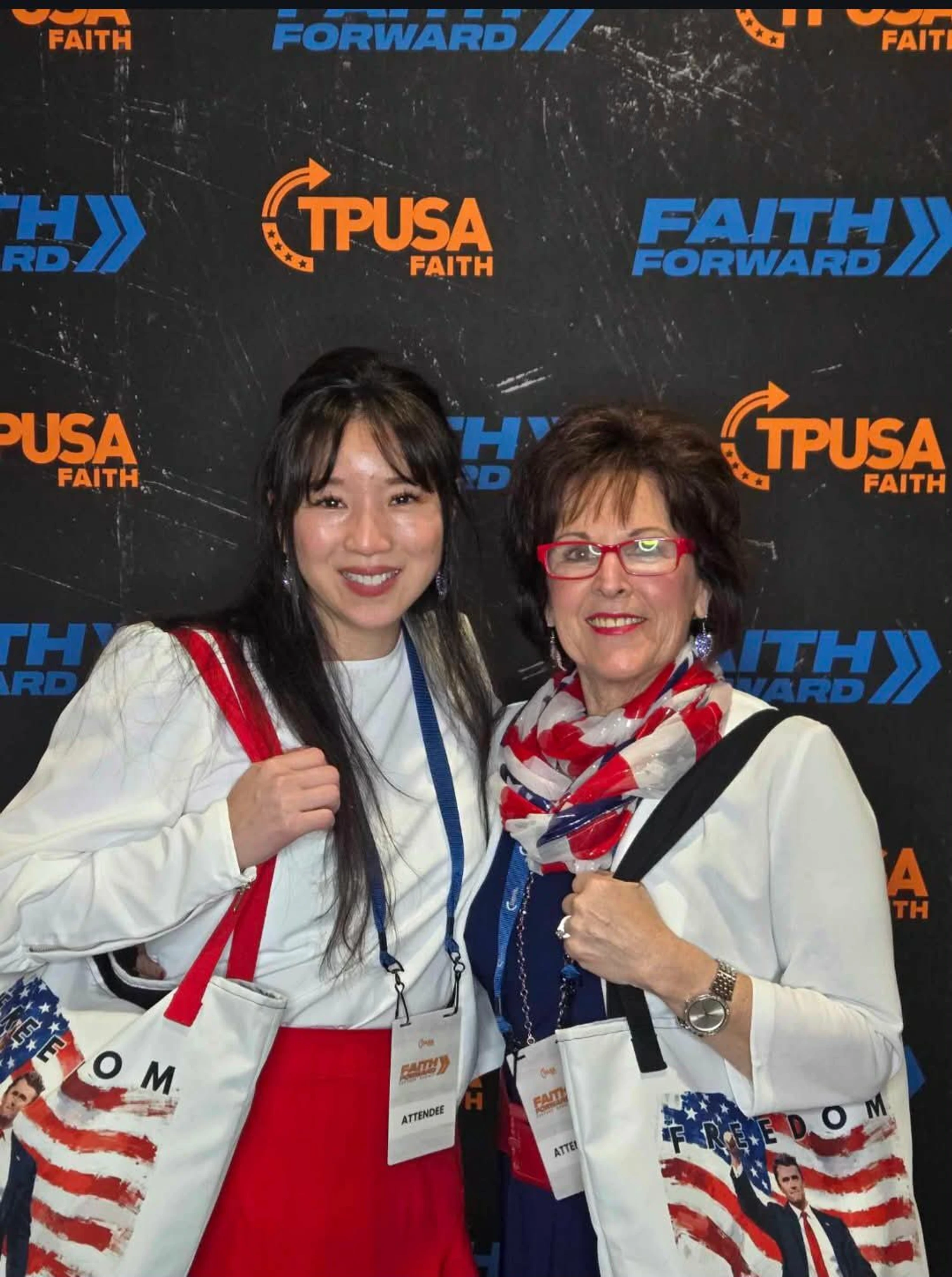 Two smiling women pose in front of a TPUSA Faith Forward event backdrop. They are both wearing attendee lanyards and carrying patriotic tote bags.