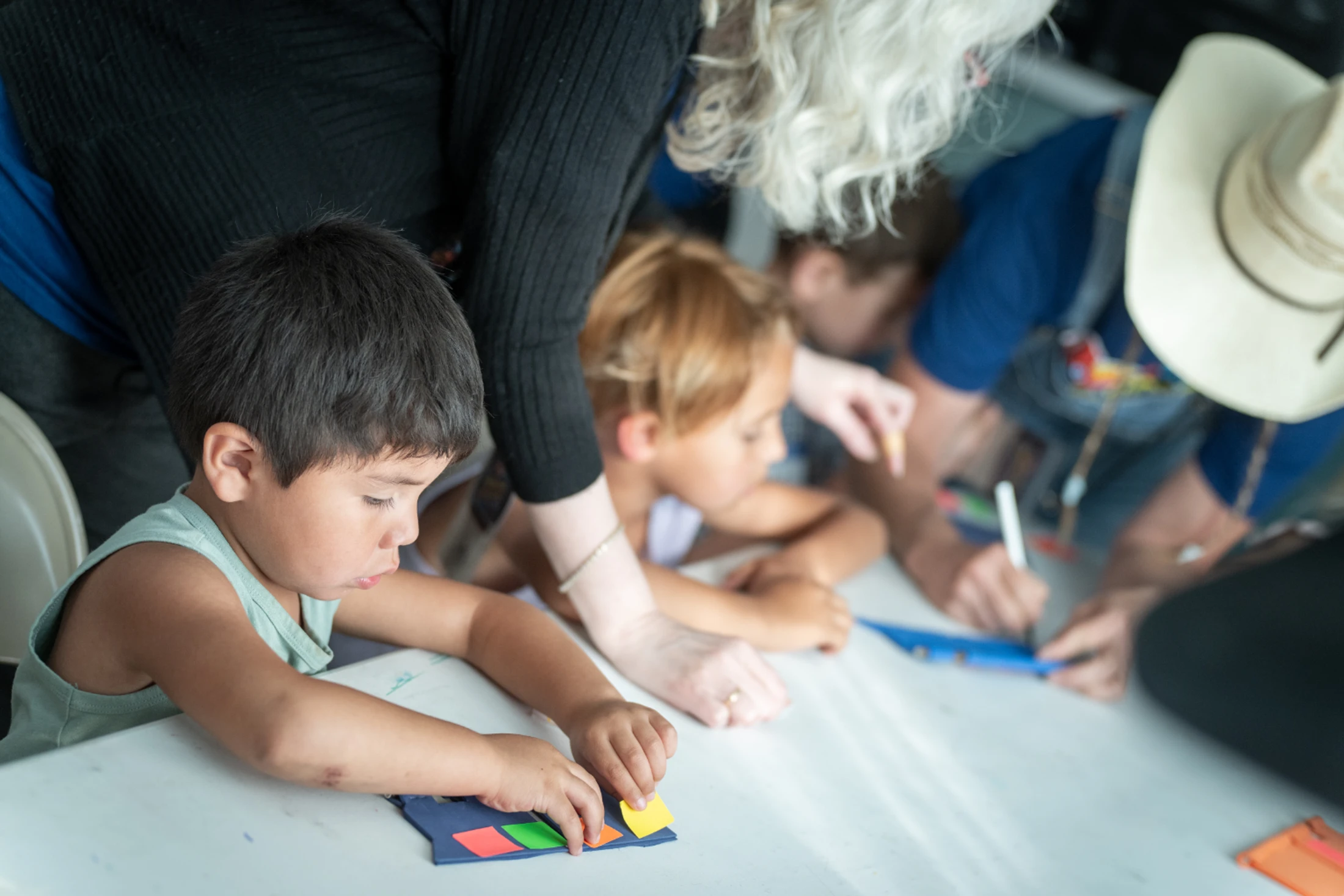 A young boy concentrates on an arts and crafts project at a table, surrounded by other children and adults who are also participating in the activity.