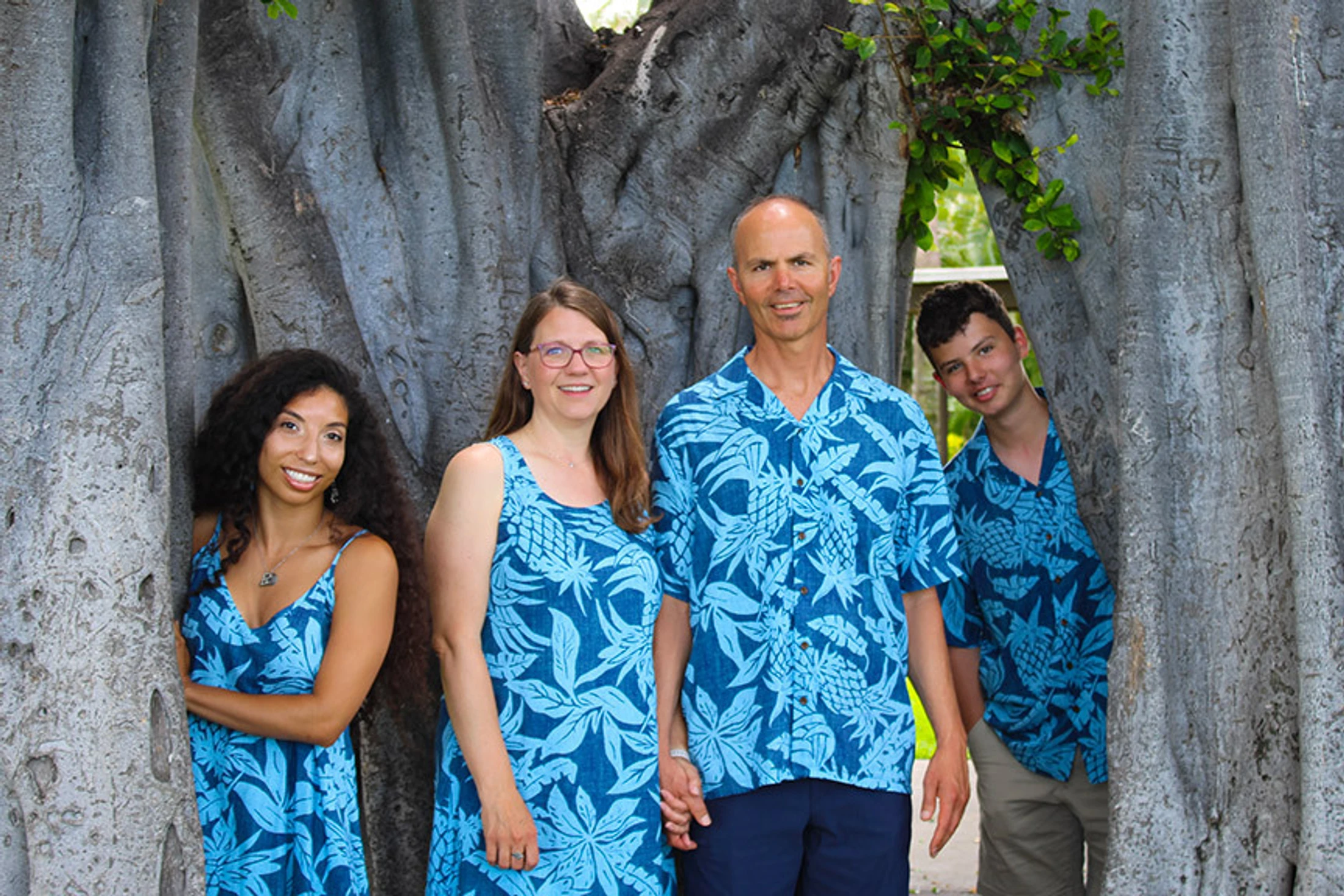 A smiling family of four in matching blue Hawaiian print outfits poses for a photo, framed by the large grey trunks of a banyan tree.