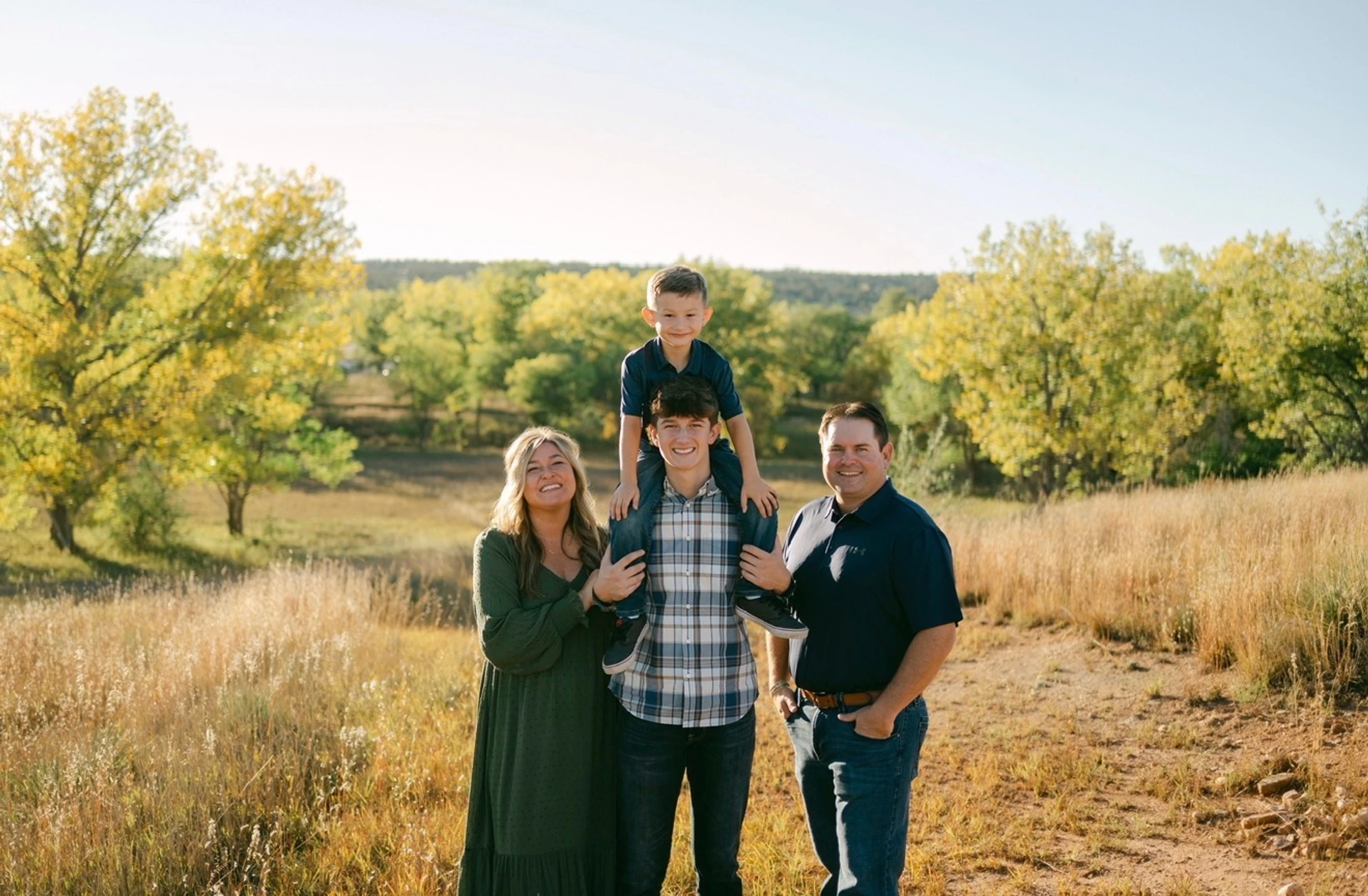 A smiling family of four stands in a sunny, grassy field. The younger son sits on his older brother's shoulders, flanked by their parents.