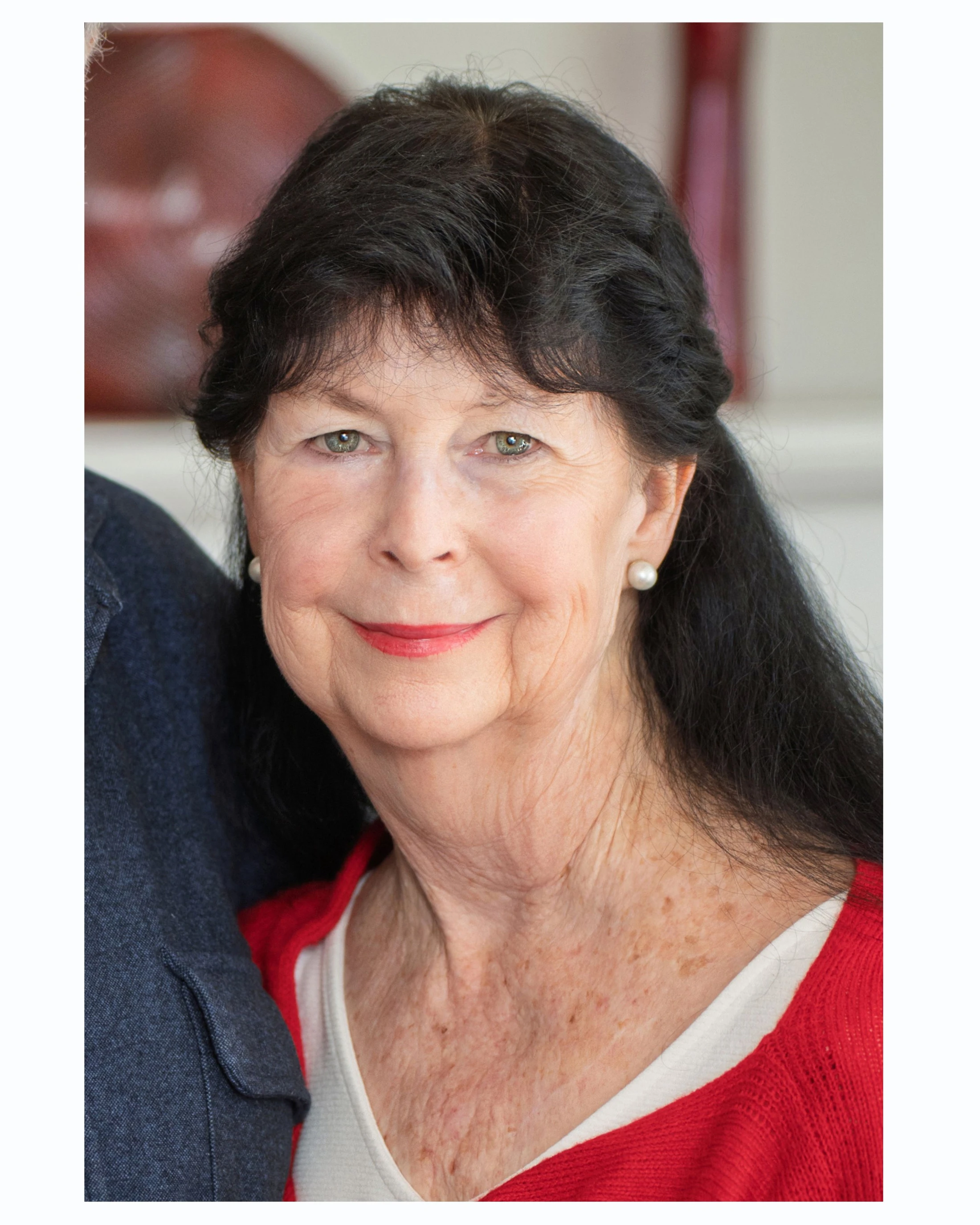A close-up headshot of an older woman with long dark hair and light eyes. She is smiling gently and wearing a red sweater over a white top, with pearl earrings.