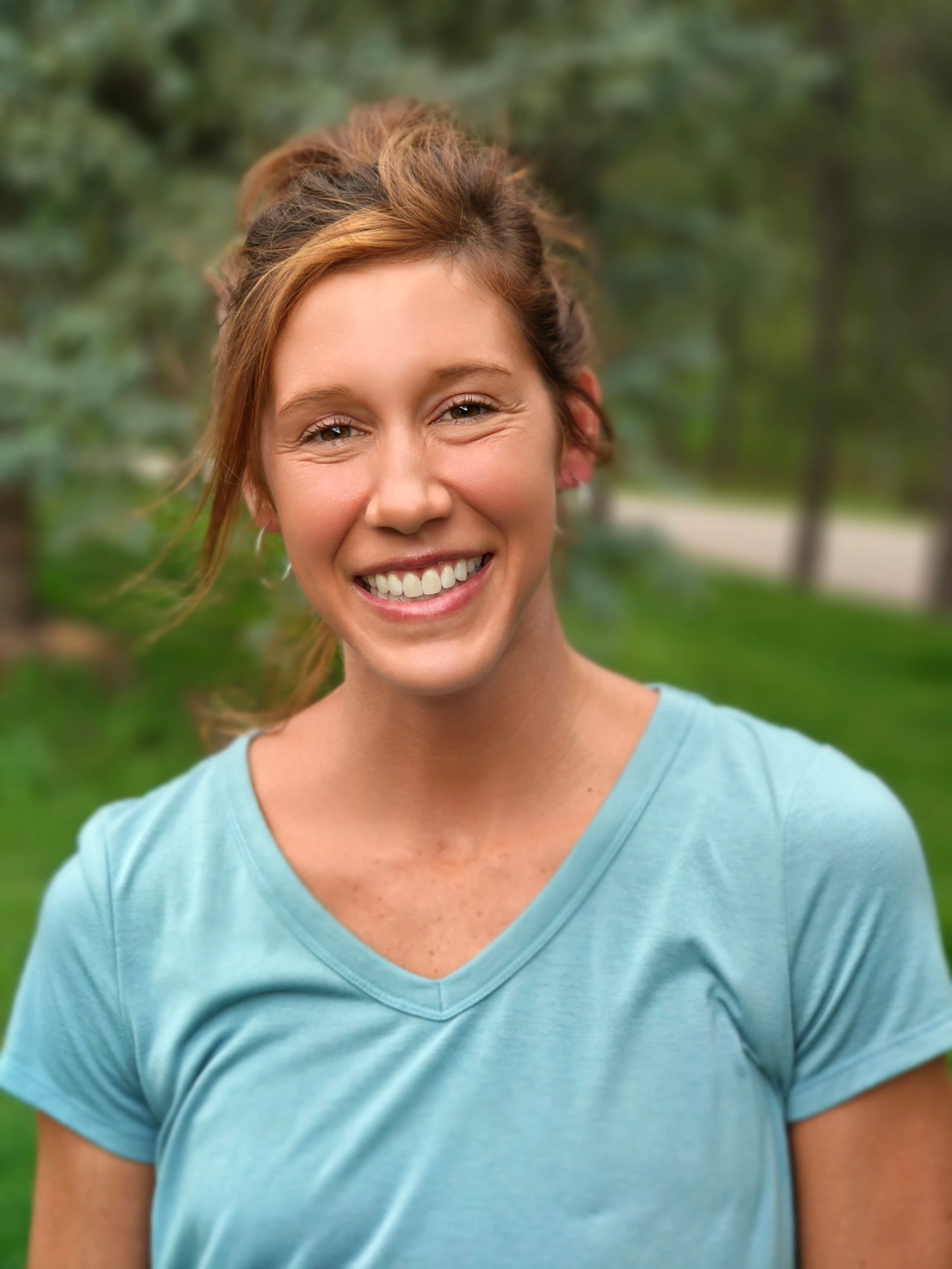 A head and shoulders portrait of a smiling woman with reddish-brown hair in an updo. She wears a light blue V-neck shirt and stands in a park.