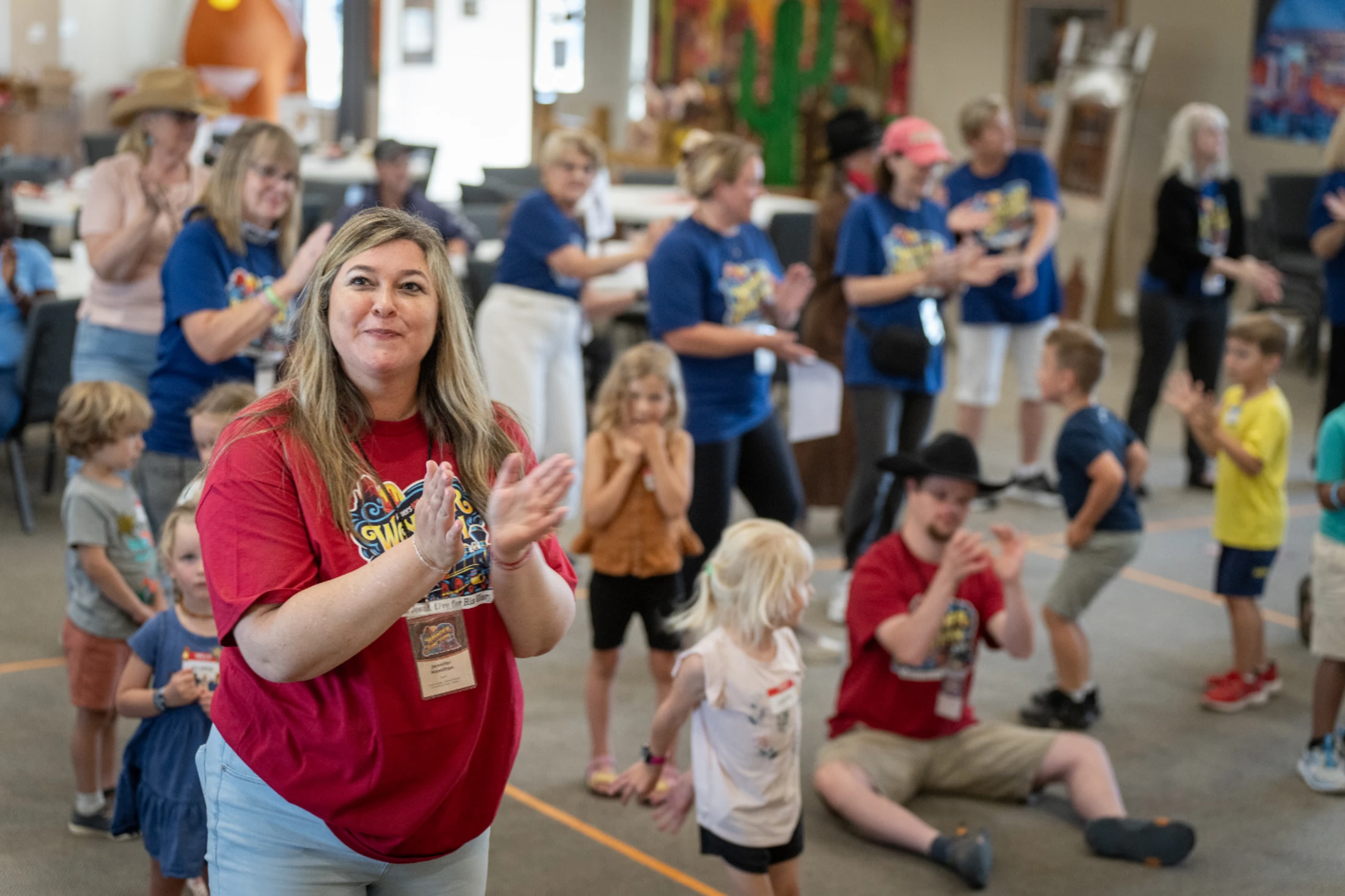 A woman in a red t-shirt smiles and claps, surrounded by other adults and children also clapping and participating in a lively indoor group activity.
