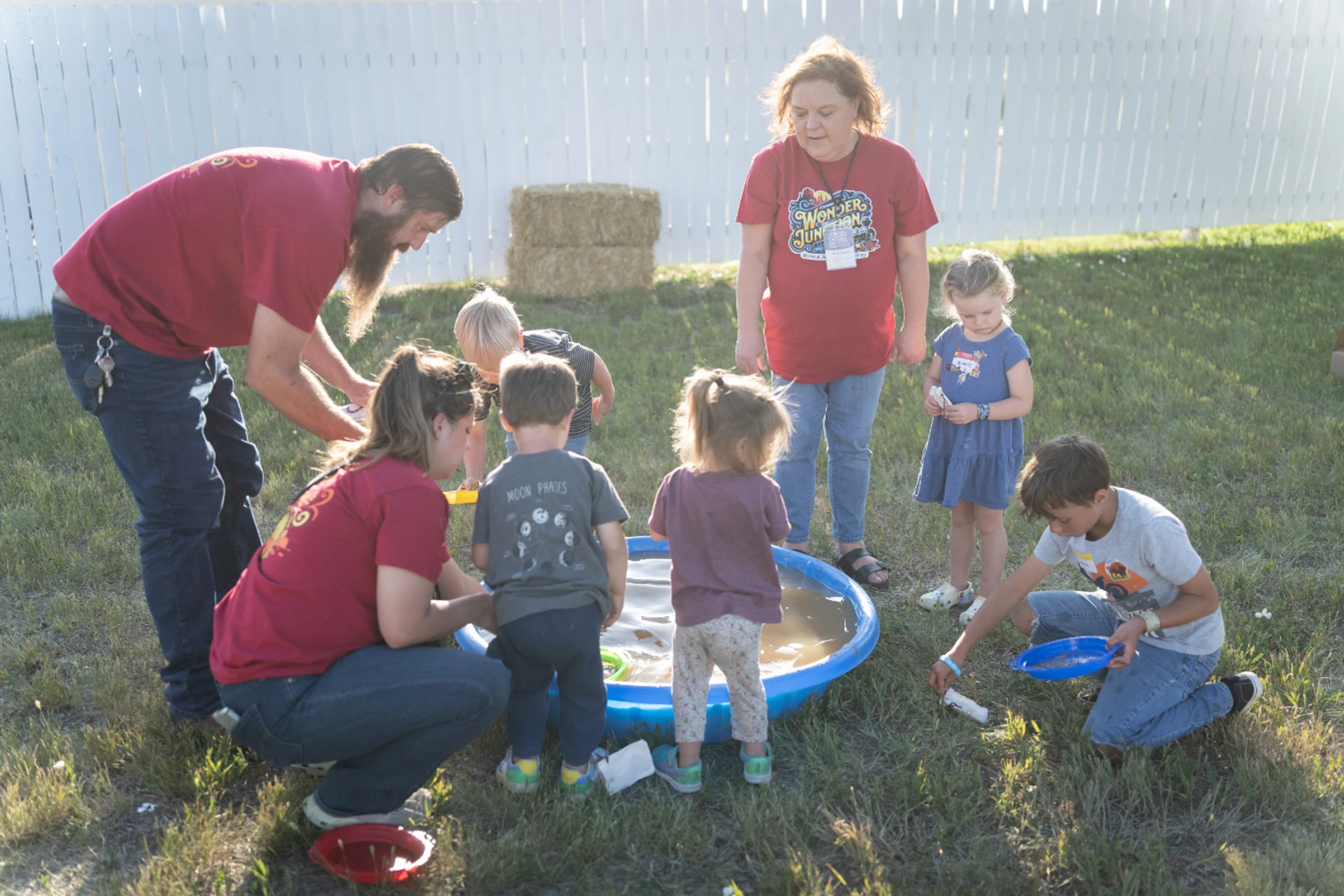 A group of adults and children gather around a small blue wading pool on a grassy lawn. The adults wear red shirts and assist the children with the activity.
