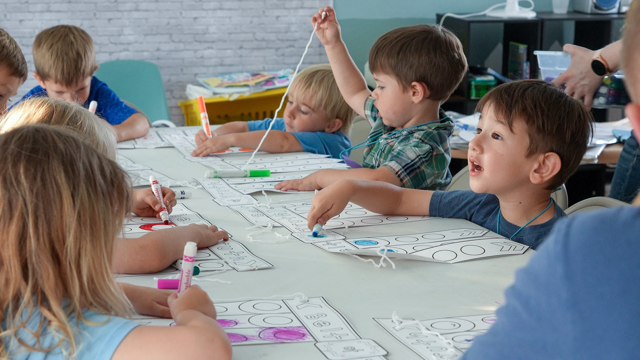 A group of young children sitting around a large white table in a classroom, focused on an arts and crafts project with markers and paper.