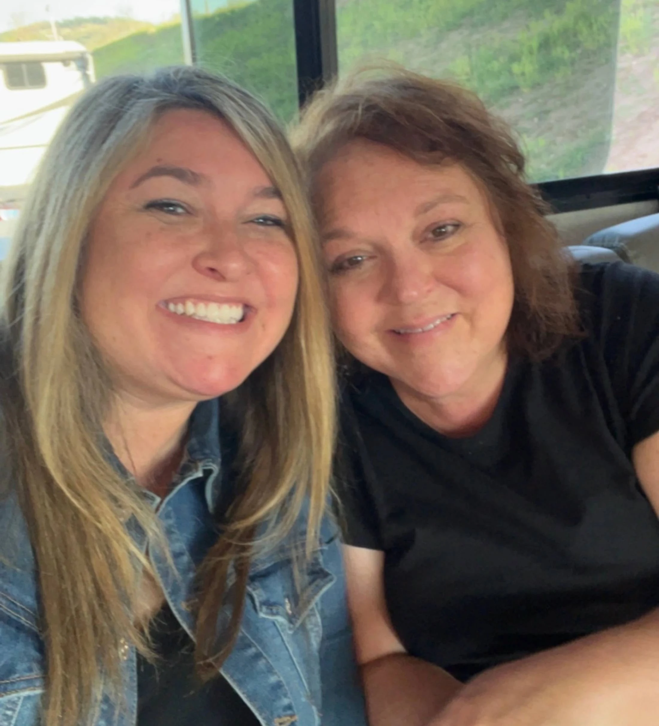 A close-up selfie of two smiling women. The woman on the left has long blonde hair and a denim jacket; the woman on the right has brown hair and a black shirt.