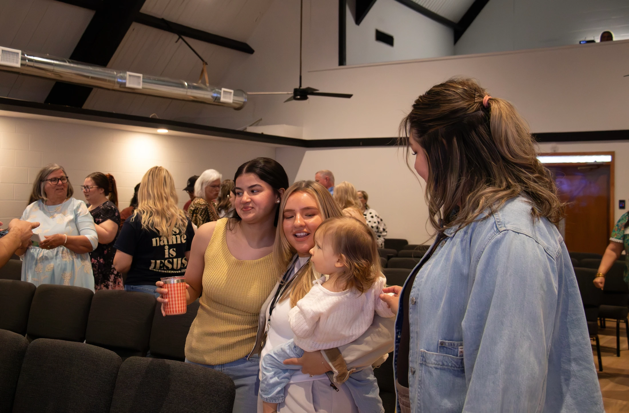 Three women gather around a baby in a church hall. One woman holds the child as they all smile and interact. Other people are in the background.