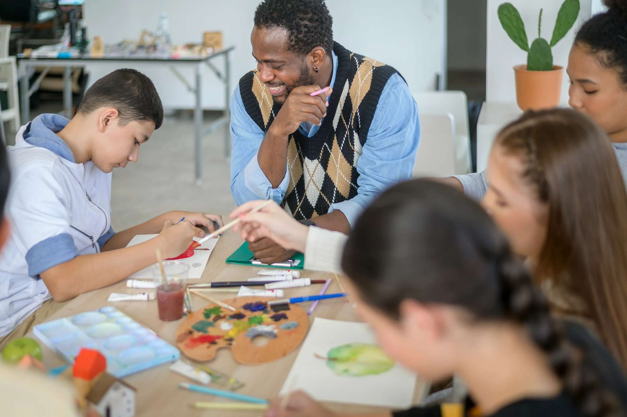 A smiling Black male teacher in an argyle sweater vest watches over a diverse group of young students as they paint at a table filled with art supplies.