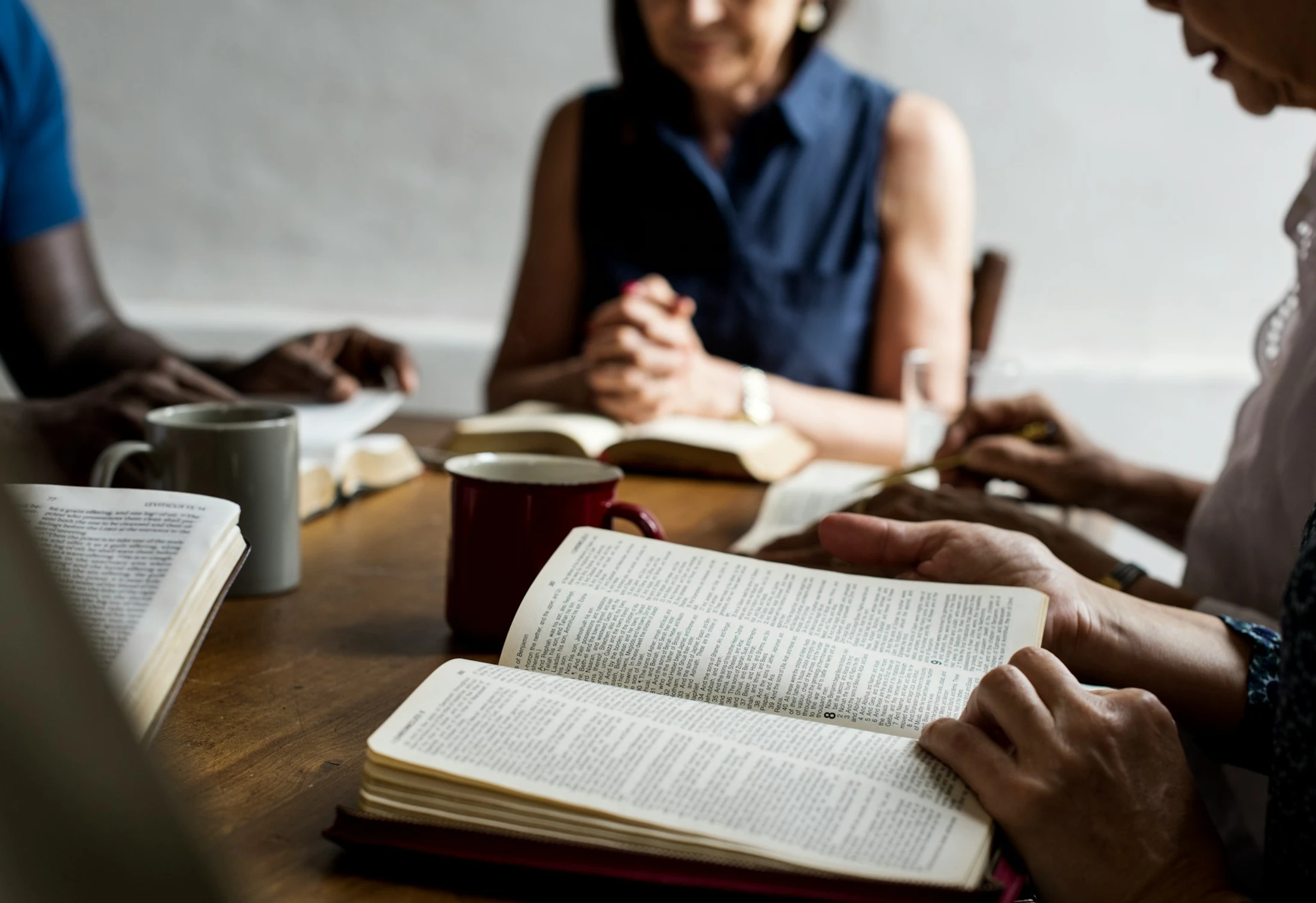 A diverse group of people sit around a wooden table reading from Bibles. The focus is on a pair of hands holding an open book in the foreground.