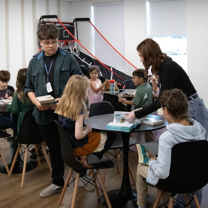 A group of children sit around tables with books as two young adult volunteers help them. A mini basketball hoop is visible in the background.