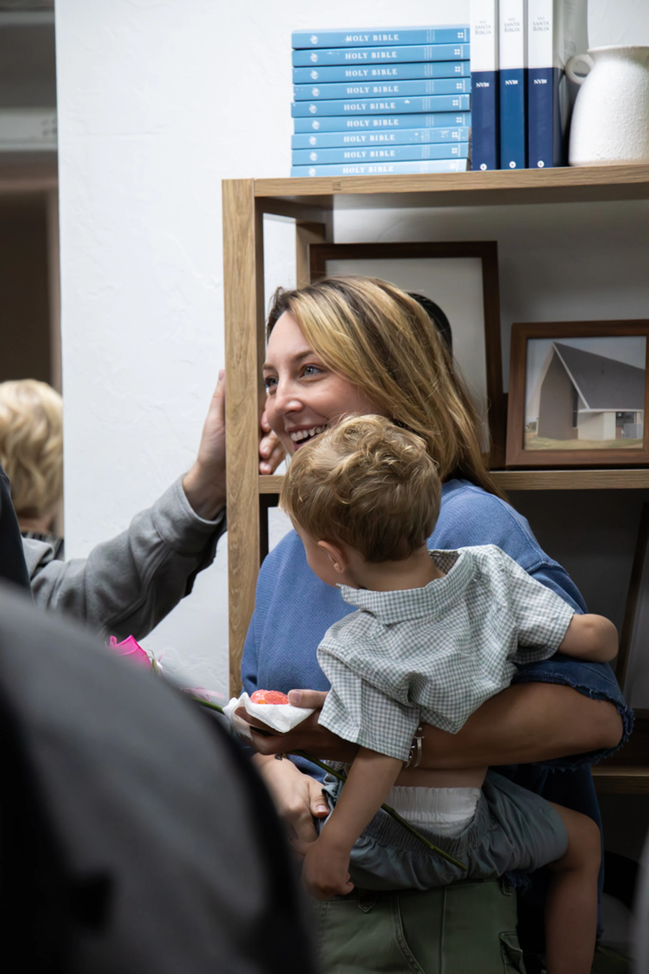 A smiling woman with blonde hair holds a toddler on her hip. In the background is a wooden bookshelf with a stack of Bibles on the top shelf.