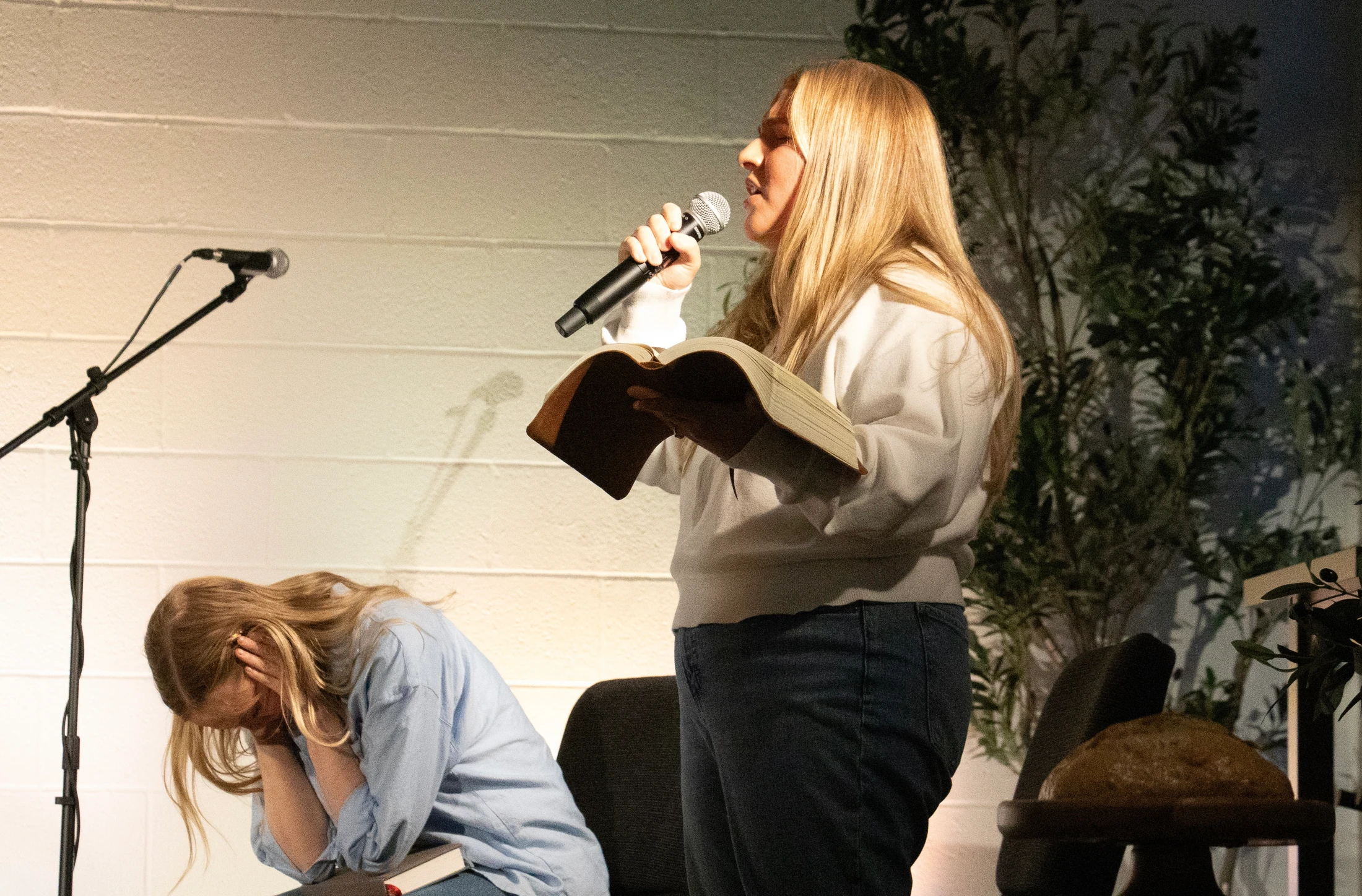 A woman stands holding a Bible and speaking into a microphone while another woman sits in front of her with her head bowed in prayer.
