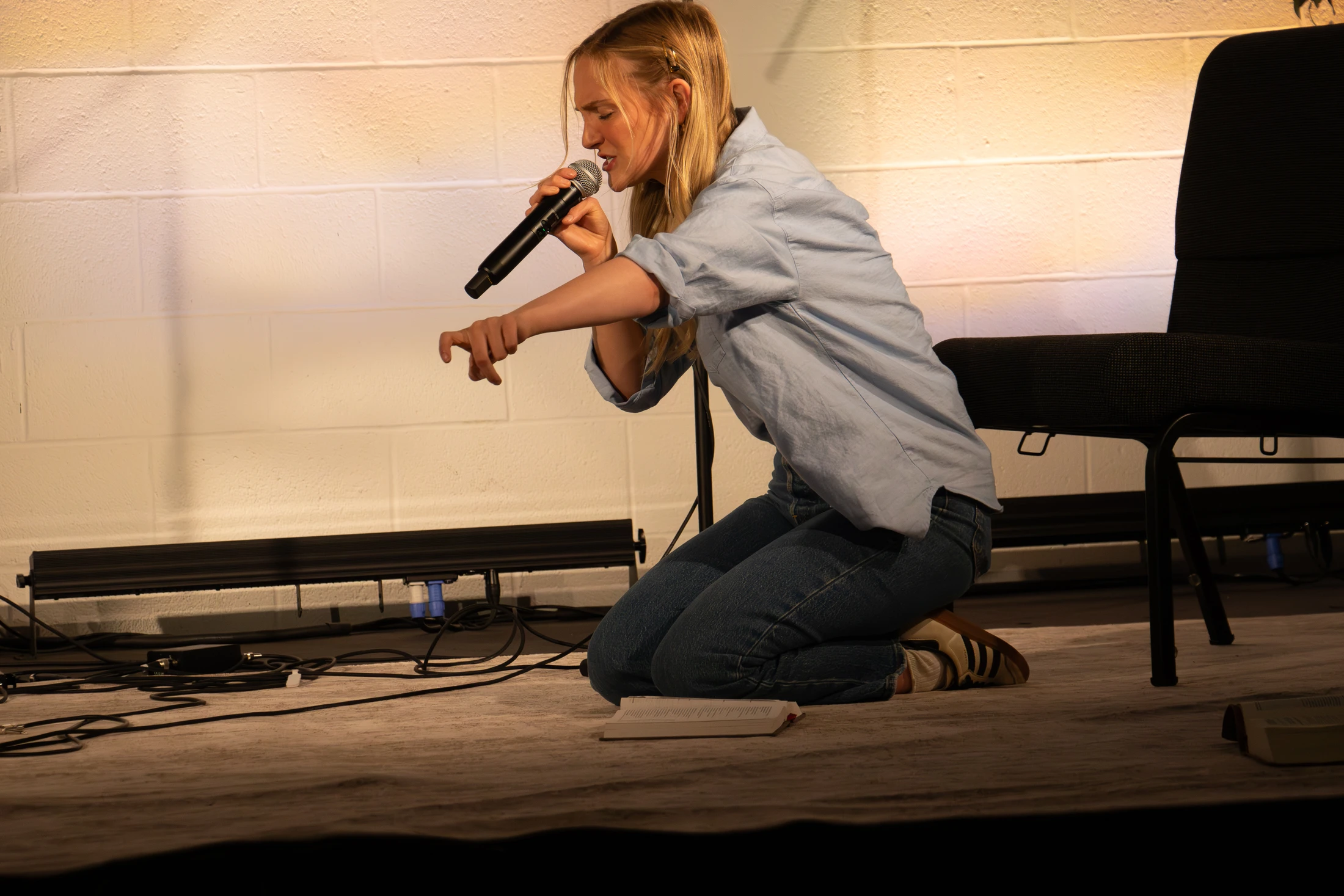 A young woman with blonde hair kneels on a stage, speaking into a microphone with her eyes closed and gesturing with her hand.