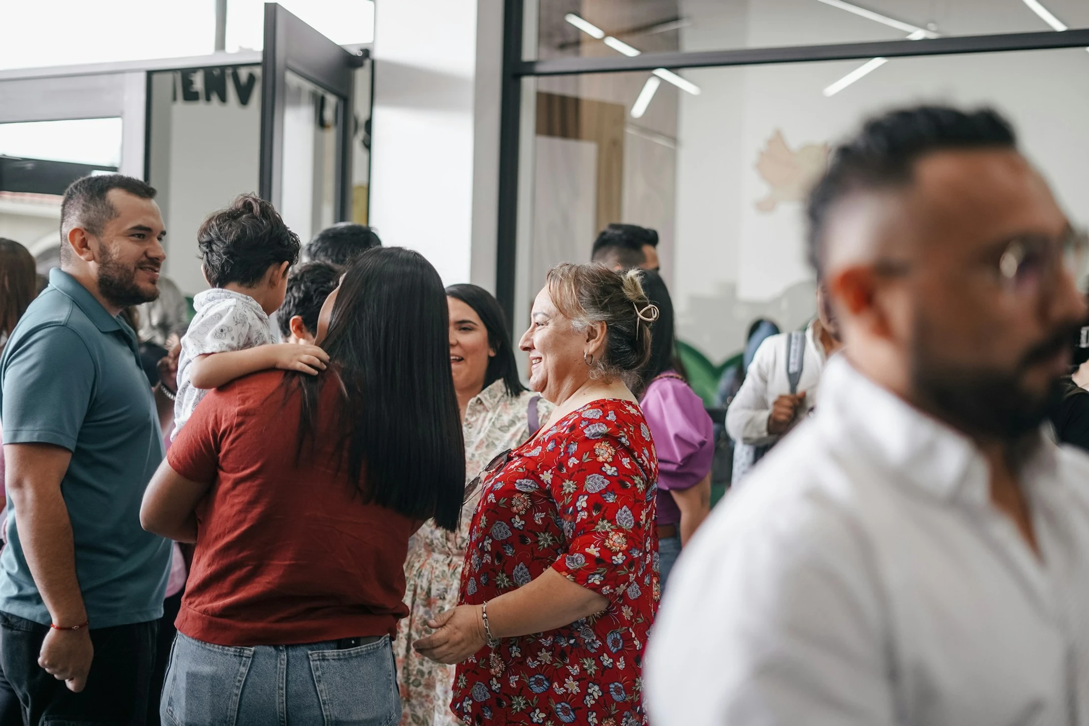 A diverse group of people stand together in a brightly lit room, smiling and talking. A woman holds a small child while another woman smiles warmly.
