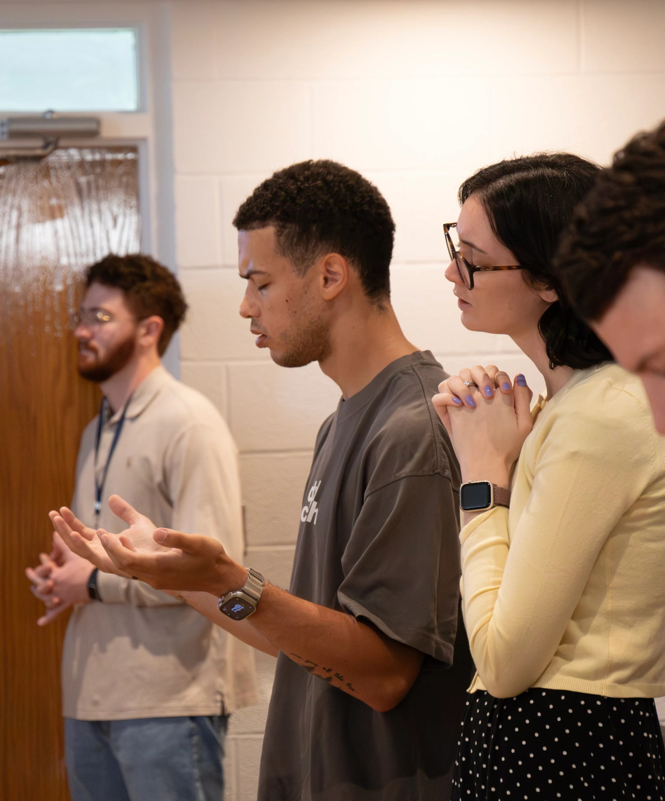 A diverse group of young adults stands side-by-side with eyes closed in prayer. A man holds his palms up while a woman next to him clasps her hands.