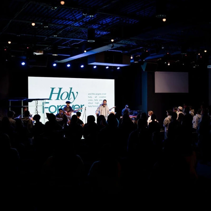 A silhouetted audience watches a worship band perform on a blue-lit stage, with a screen displaying the words 'Holy Forever' behind the lead singer.
