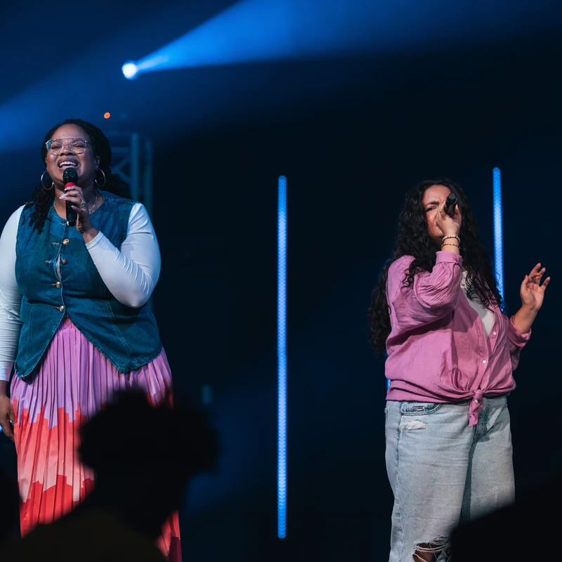 Two women sing into microphones on a dark stage with blue spotlights. The woman on the left smiles, wearing a colorful skirt, while the other sings emotionally.