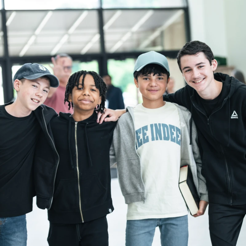 Four diverse young boys in casual clothing stand with their arms around each other, smiling for the camera in a brightly lit building lobby.