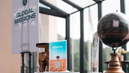 A table displays materials for a global missions event, including flyers, a small globe, and a wooden elephant, set against a bright window.