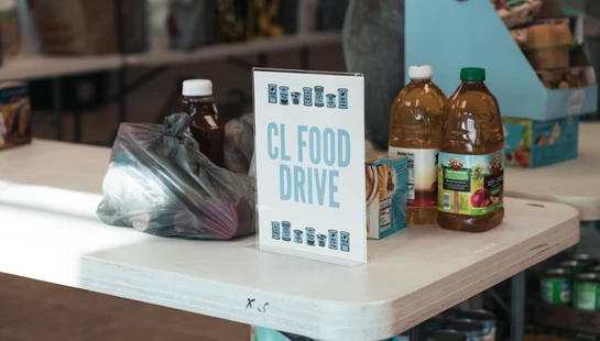 A sign that reads 'CL Food Drive' on a white table surrounded by donated groceries like juice, canned goods, and items in plastic bags.