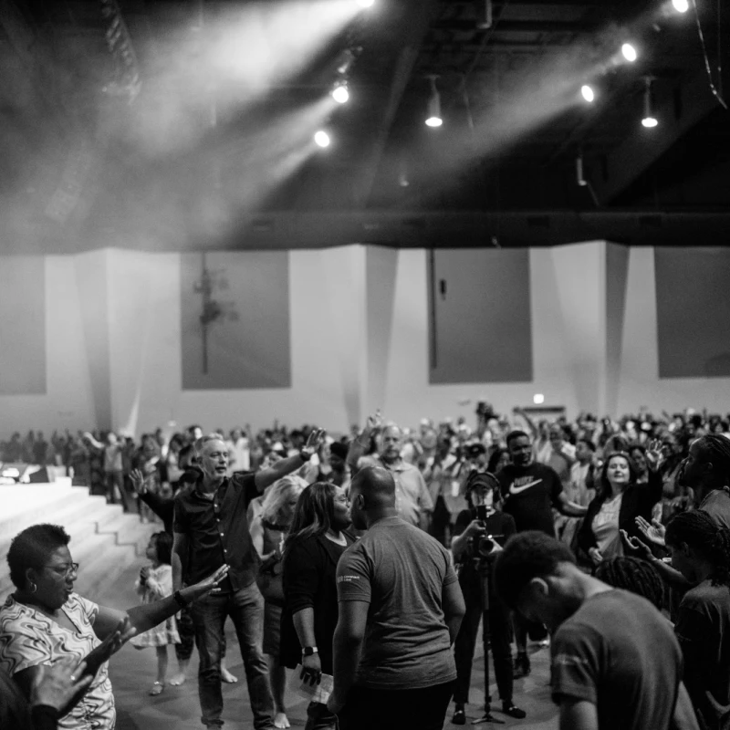 A black and white photograph of a large, diverse crowd in a hall, with many people raising their hands in worship during an event with a stage and lights.