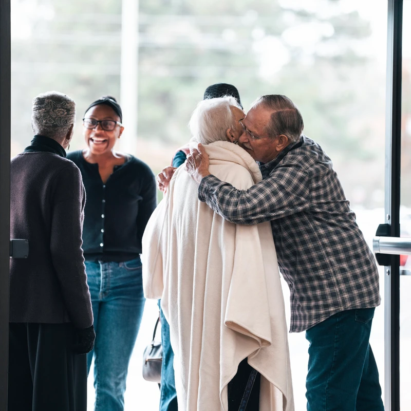 An elderly man in a plaid shirt hugs and kisses an elderly woman in a cream shawl at a doorway, while two other women watch and smile warmly.