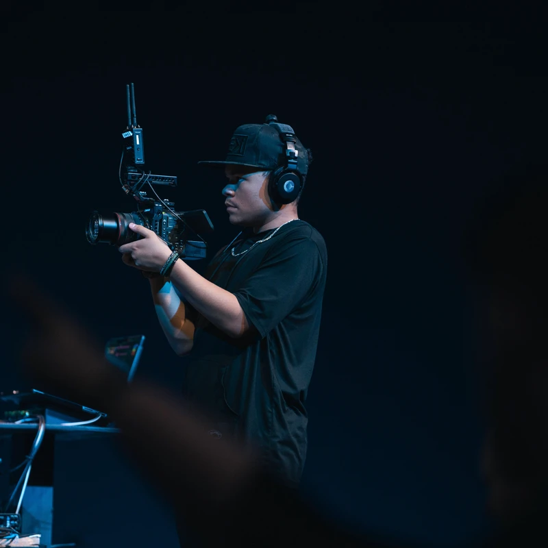 A young man wearing a cap and headphones films with a professional video camera rig against a dark background with blue lighting.