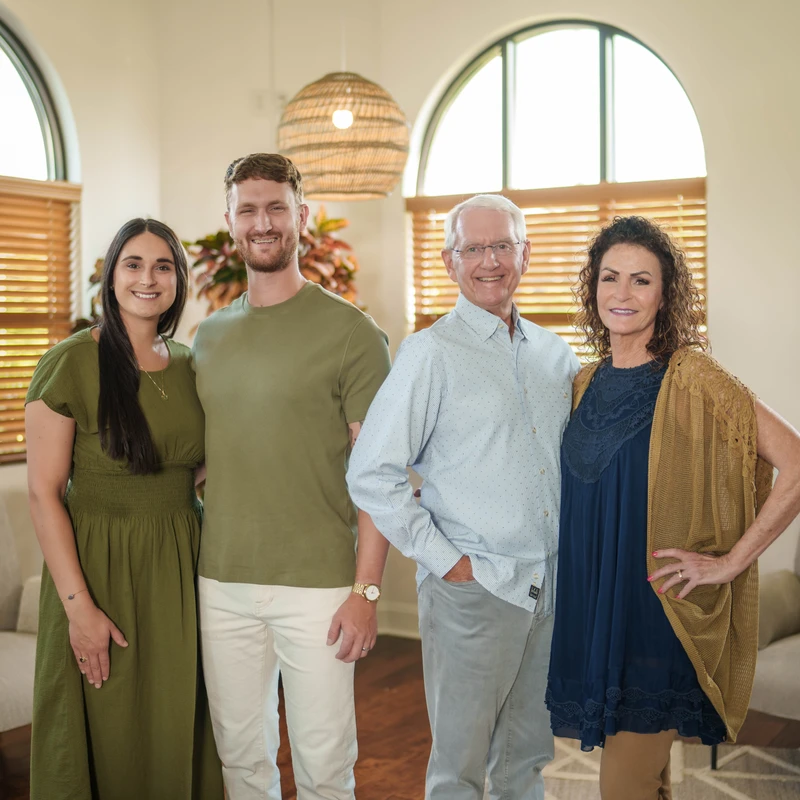 A family of four, an older couple and a younger couple, stand together smiling for a portrait in their modern, brightly lit living room.