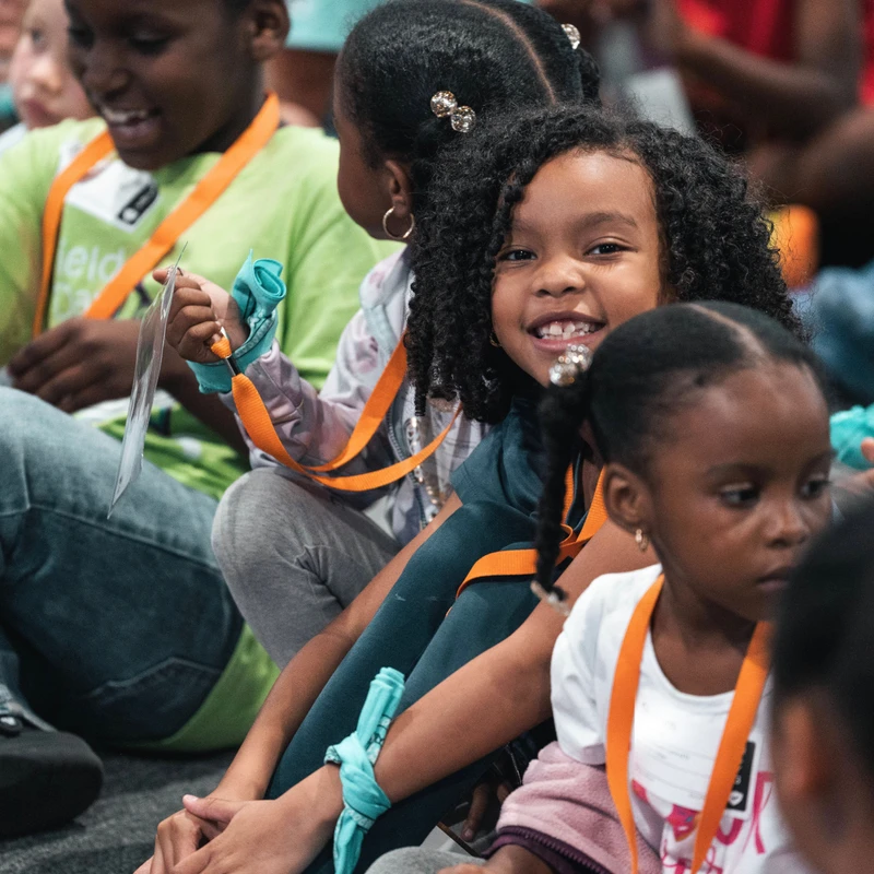 A group of young Black children sit together on the floor. One girl with curly hair smiles warmly at the camera, while others around her are also engaged.
