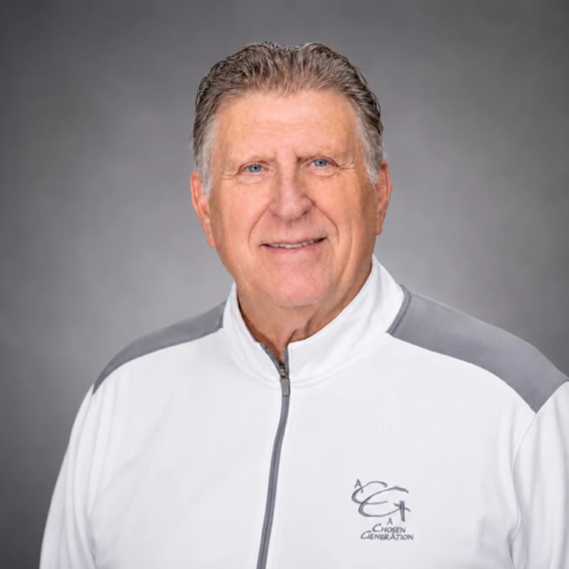A smiling older man with blue eyes and short grey hair poses for a headshot. He is wearing a white and grey zip-up jacket against a solid grey background.