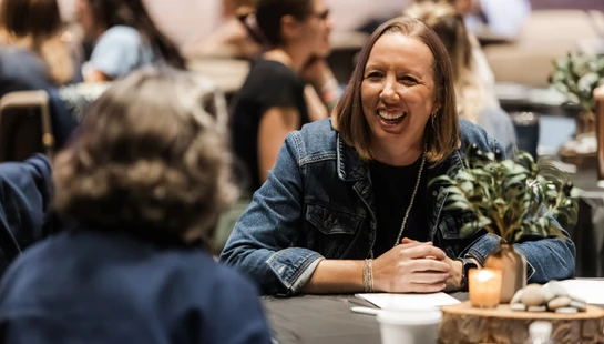 A woman with shoulder-length brown hair in a denim jacket laughs while talking to another person across a table at a gathering.