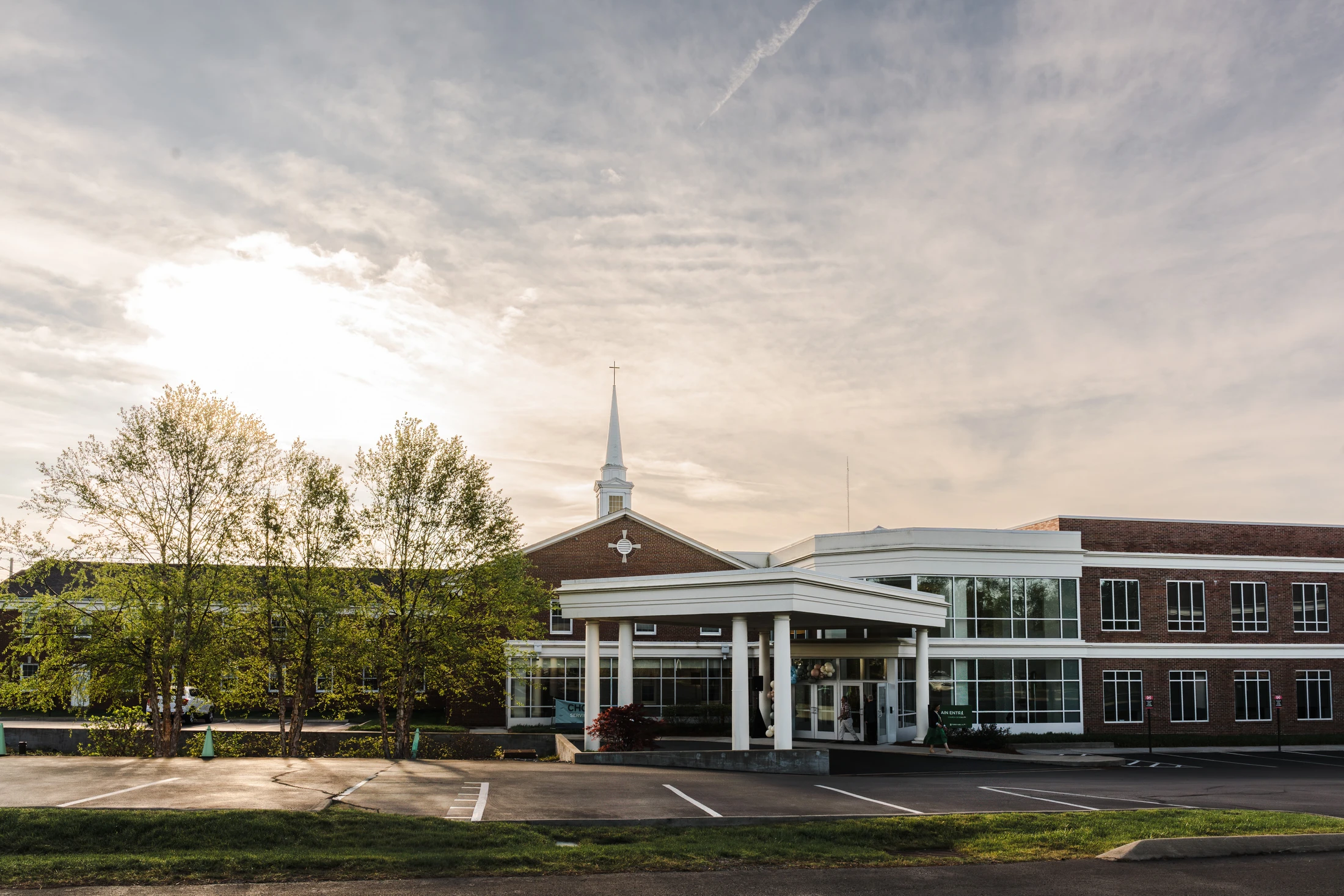 A large, modern brick church with a white steeple and a covered entrance, seen from its parking lot during a cloudy sunset.