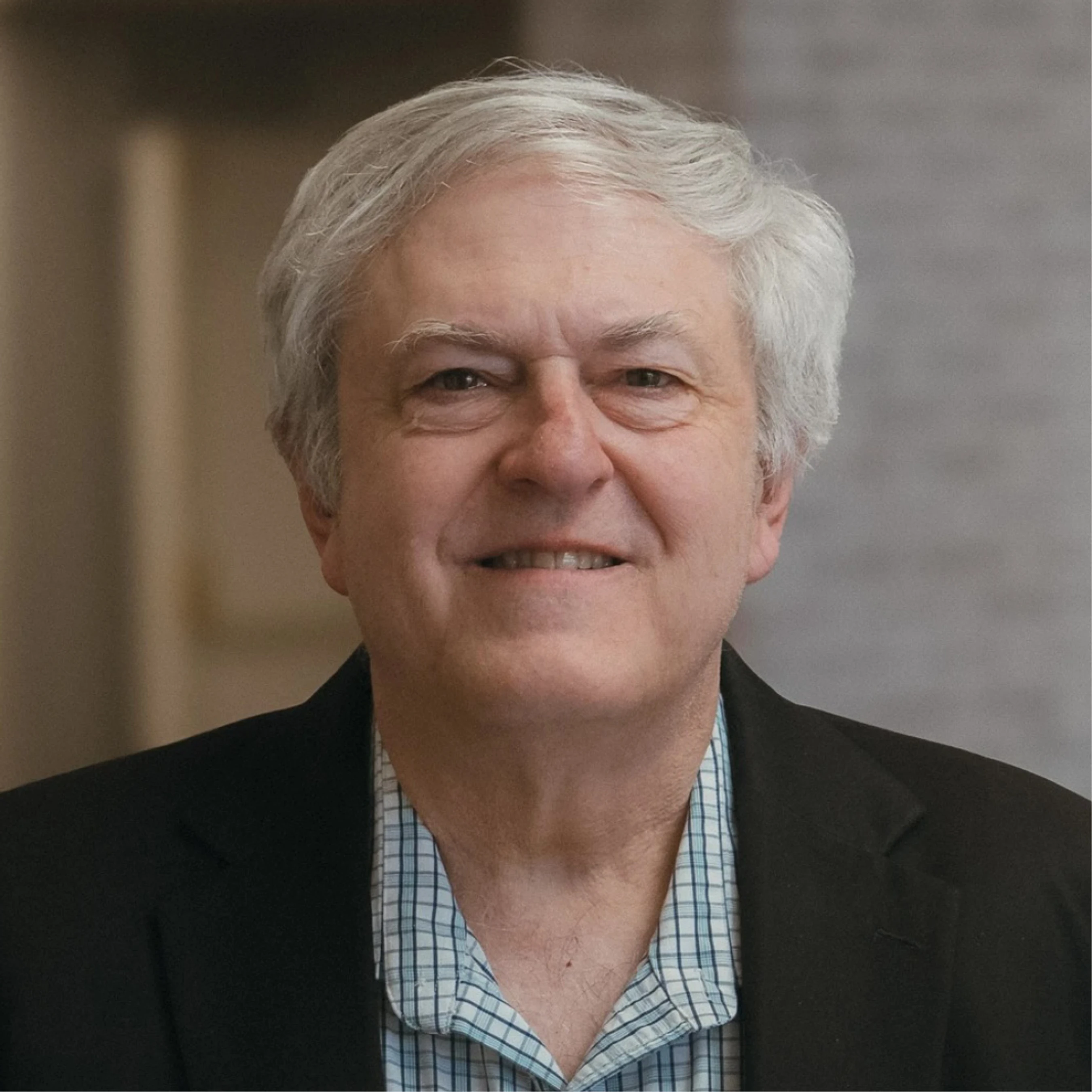 A head-and-shoulders portrait of a smiling older man with gray hair. He wears a black jacket over a checkered shirt and looks directly at the camera.