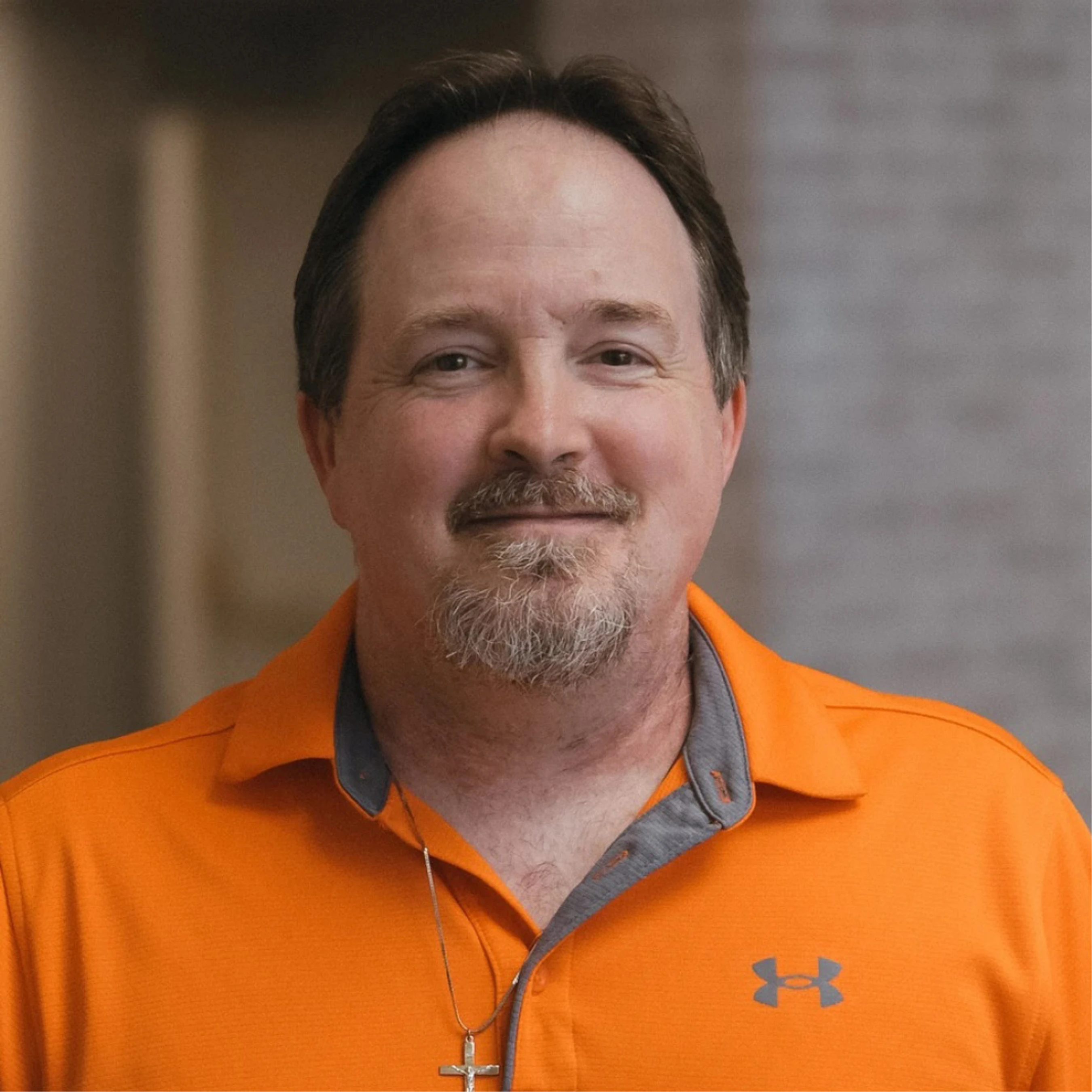 A friendly, middle-aged man with a goatee smiles for a headshot. He wears a bright orange polo shirt with a grey collar and a silver cross necklace.