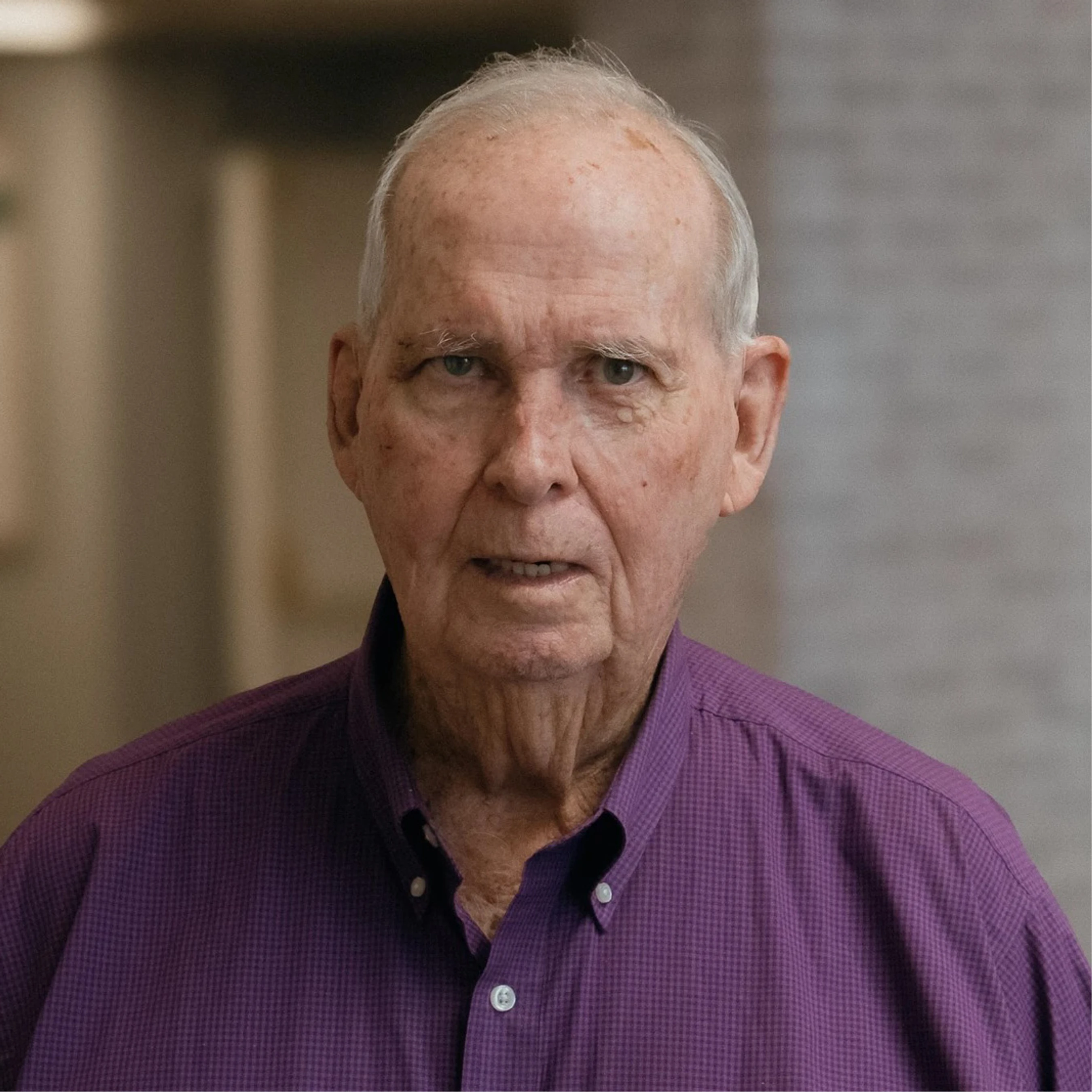 A close-up portrait of an older man with white hair wearing a purple button-down shirt. He is looking directly at the camera with a neutral expression.