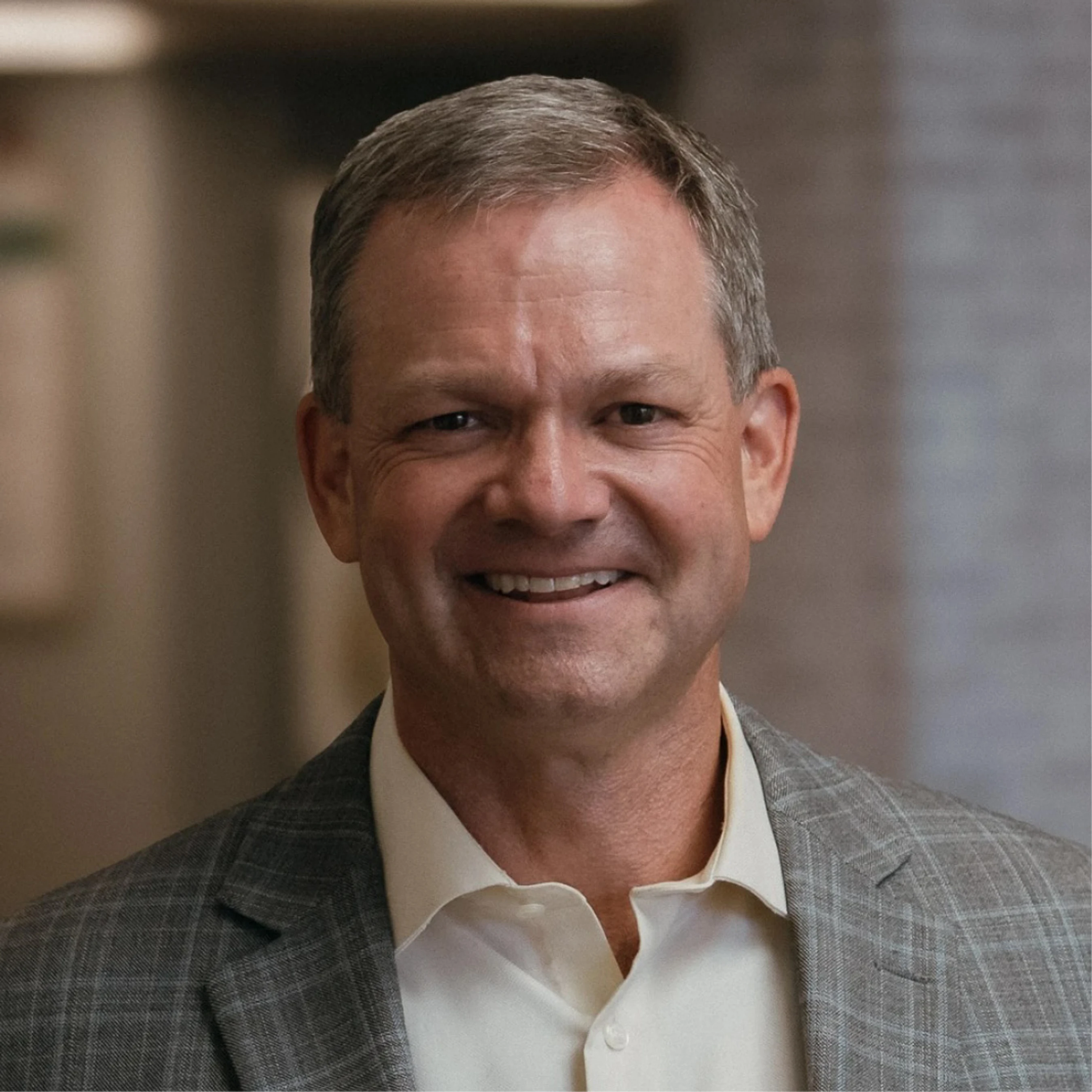 A head-and-shoulders portrait of a smiling, middle-aged white man with short graying hair, wearing a gray plaid blazer over a light-colored collared shirt.