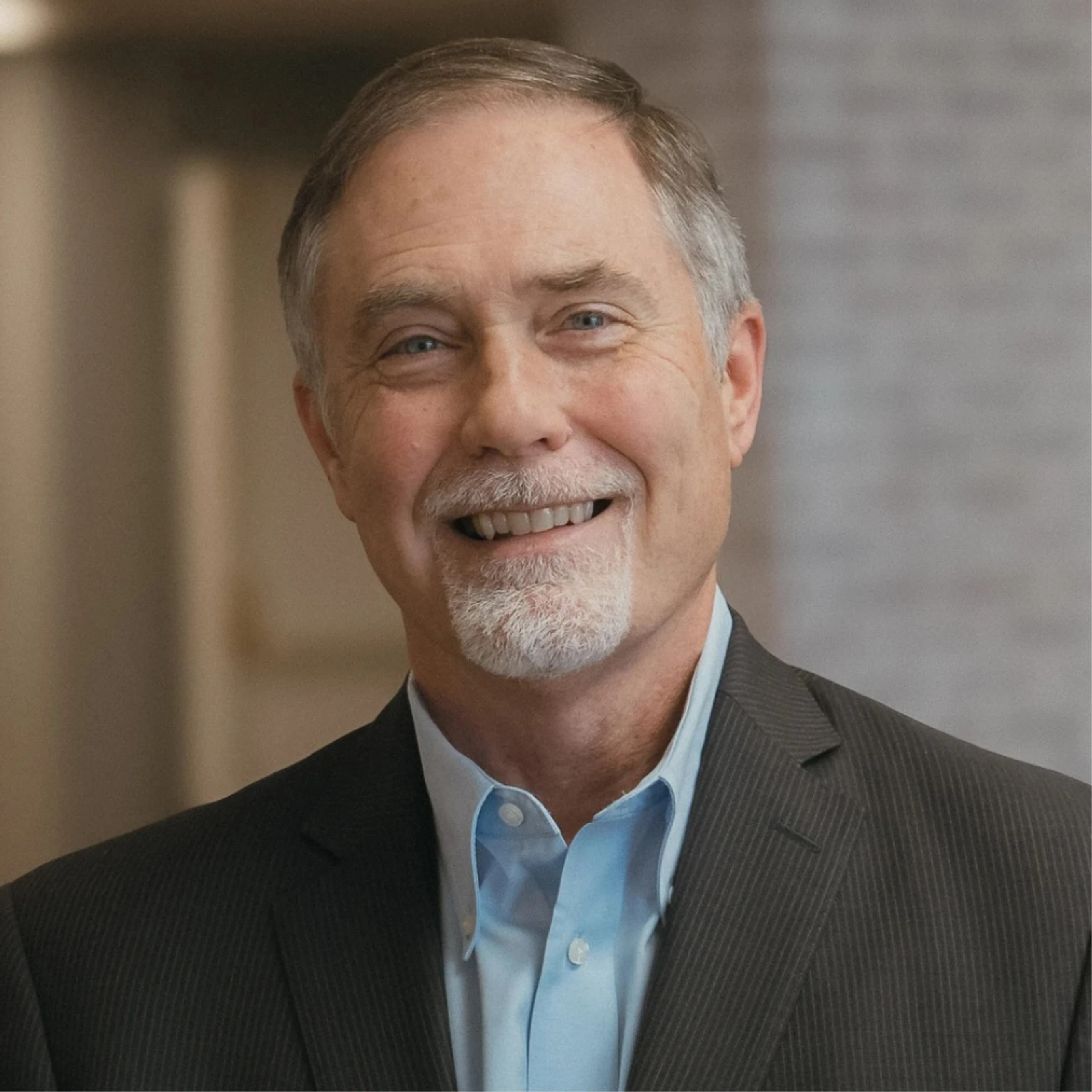 A smiling, middle-aged man with blue eyes and a gray goatee poses for a professional headshot. He wears a blue collared shirt and a dark pinstripe suit jacket.