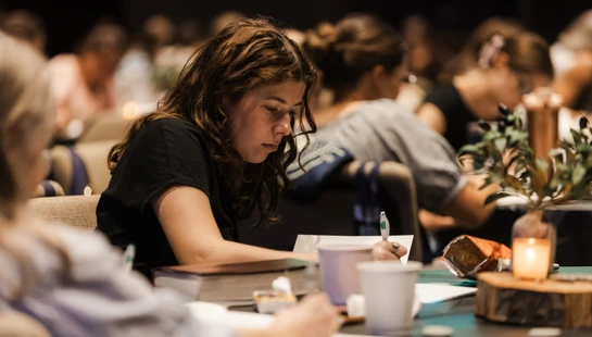 A young woman with curly brown hair wearing a black shirt concentrates as she writes notes in a notebook at a seminar, surrounded by other attendees.
