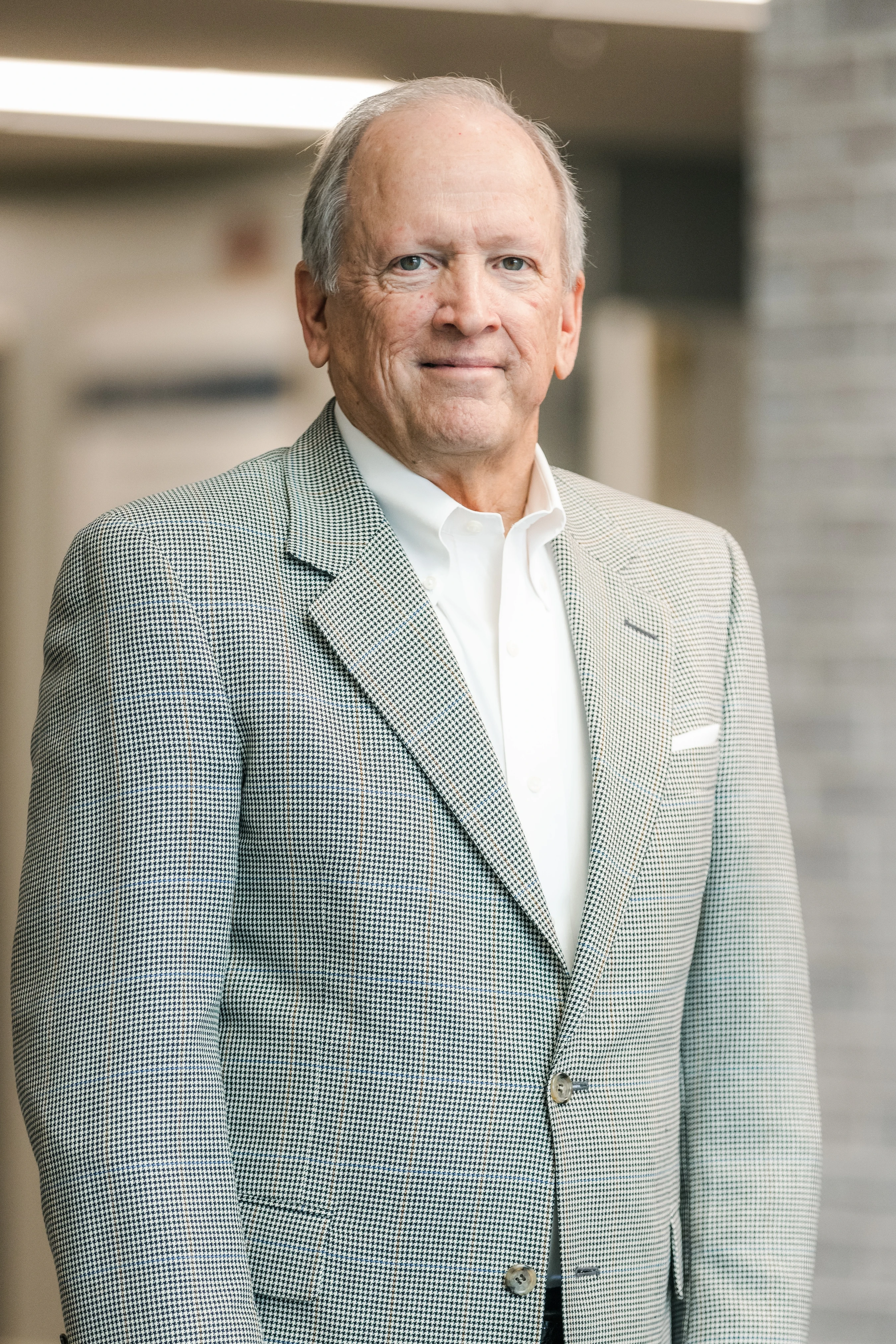 A waist-up portrait of an older man with gray hair, smiling gently. He wears a black and white houndstooth sport coat over a white collared shirt indoors.